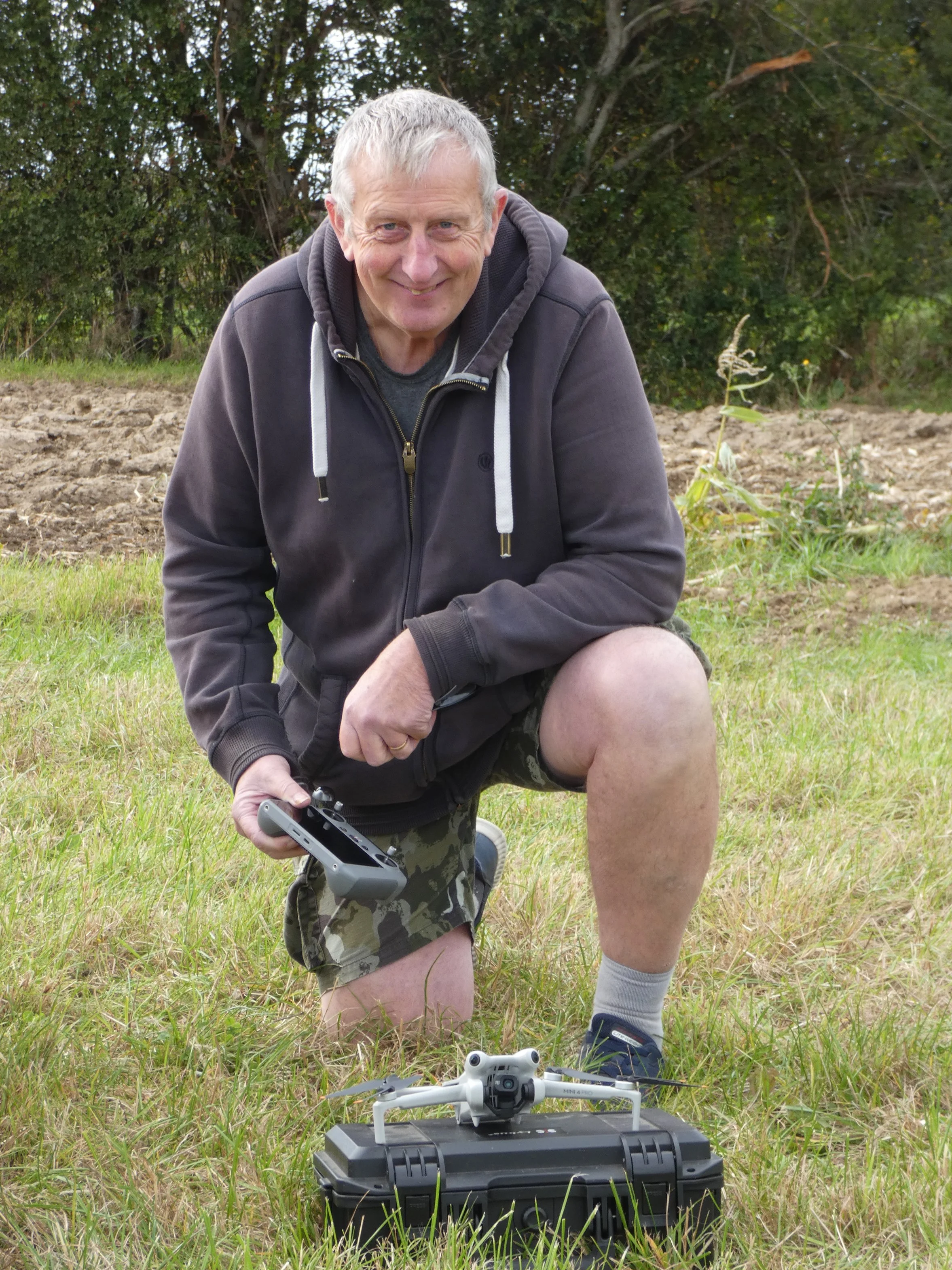 A man with gray hair smiling, squatting outdoors on grass, holding a remote control for a drone, with a white drone on a case in front of him, and a drone controller on the case, with trees and bushes in the background.