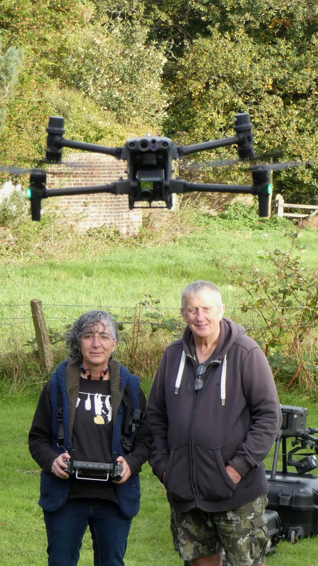 Two people standing outdoors in front of a grassy area and trees, one woman with a drone controller and the man casually dressed in camouflage shorts and a hoodie, with drone equipment nearby.