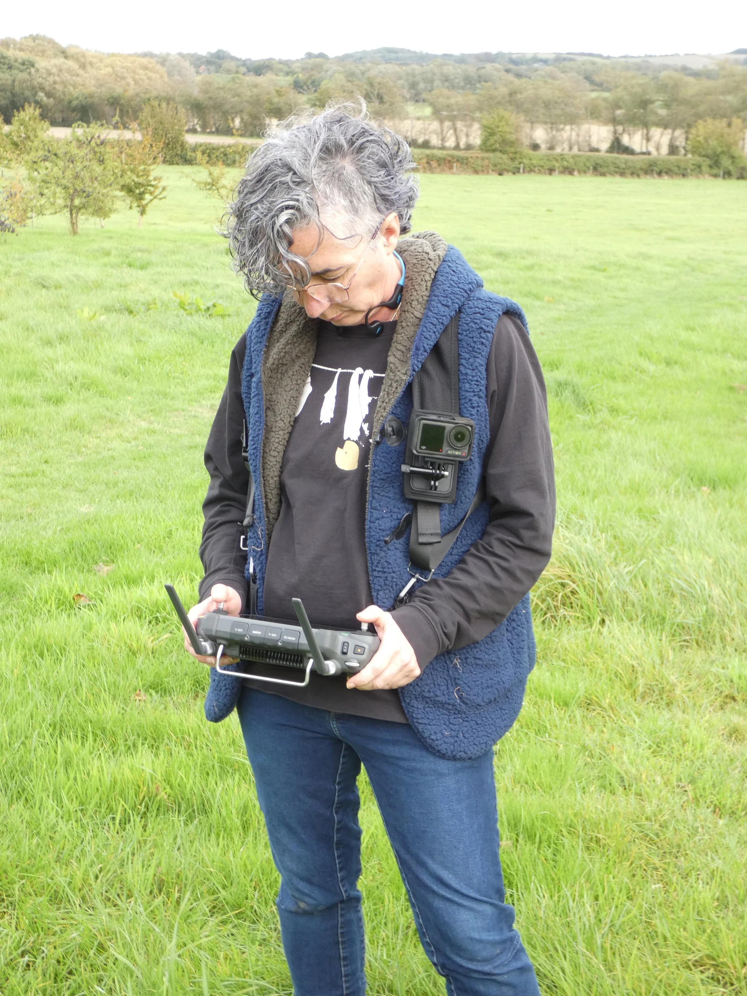 A person with gray curly hair and glasses looking down while holding a drone controller, standing on a grassy field with trees and hills in the background.