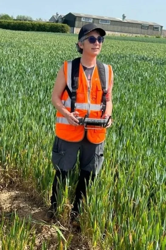 Woman in an orange safety vest and sunglasses standing in a green field, holding a device, with a house in the background.