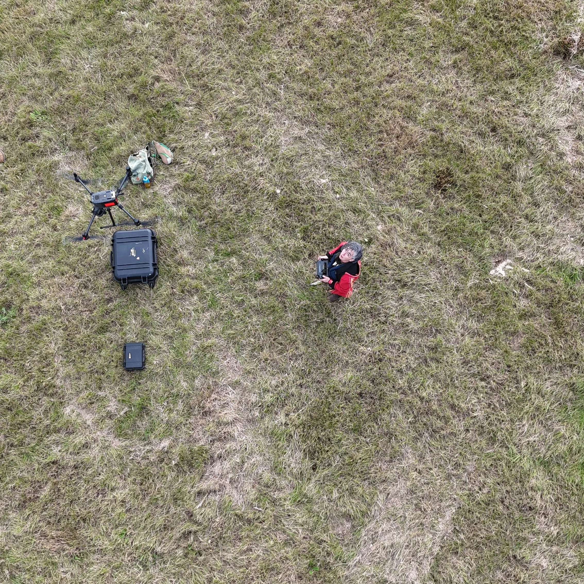Aerial view of drone operator holding a remote control, with a drone, a backpack, and a black case nearby on a grassy field.