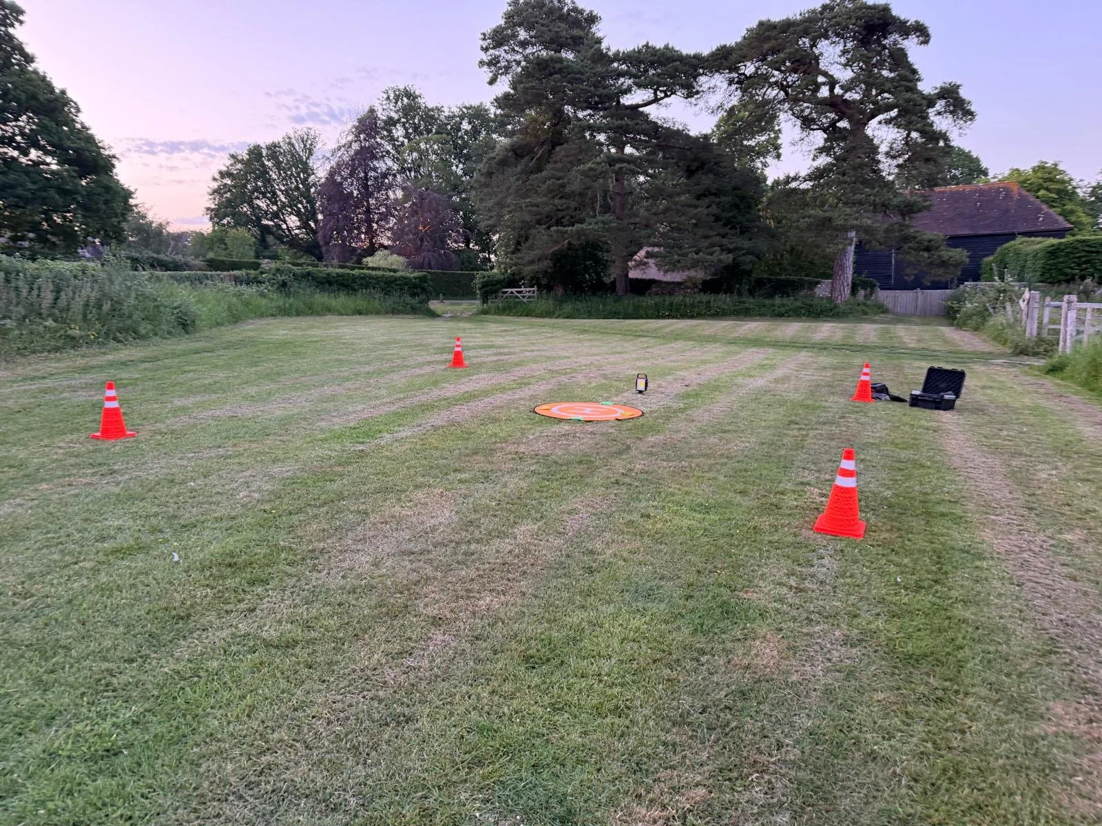 An open grassy field with four orange traffic cones surrounding a circular target and some equipment, including a small black case and a device, set up on the ground, under a cloudy sky during dusk.