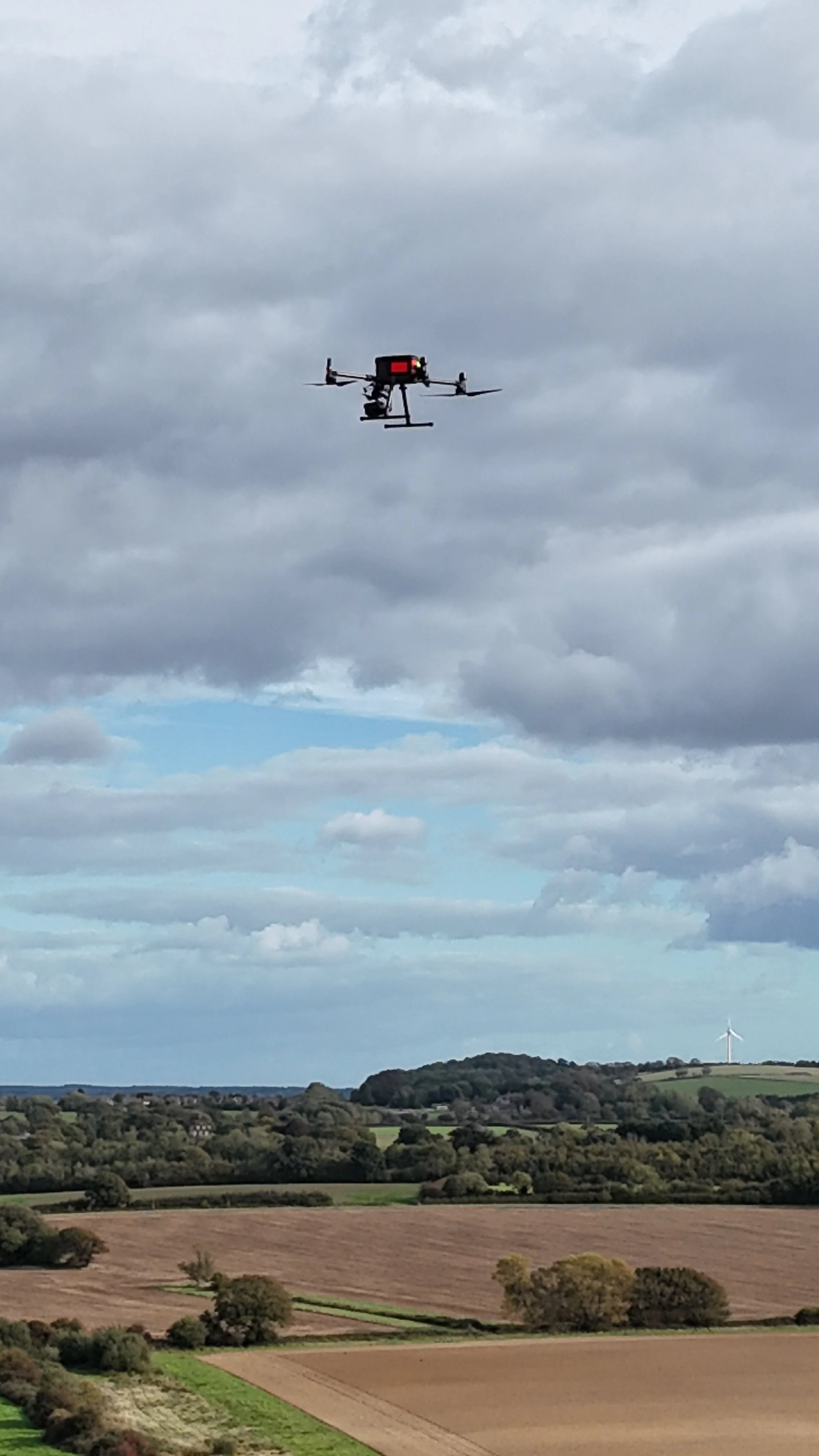 A drone flying in the sky above a rural landscape with fields, trees, and wind turbines on the horizon under cloudy weather.