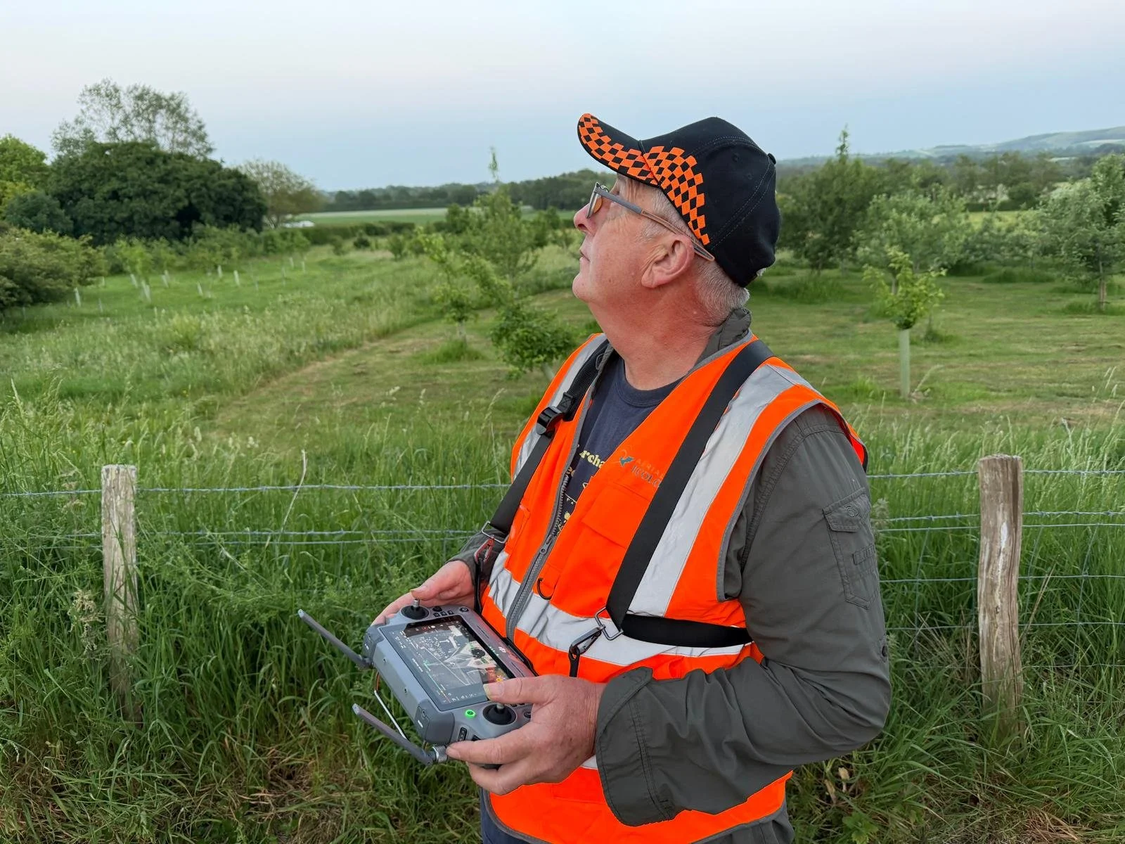 A man wearing a black cap with orange checkered pattern, orange reflective safety vest, and glasses holds a drone controller outside in a green field with trees and hills in the background, operating a drone.