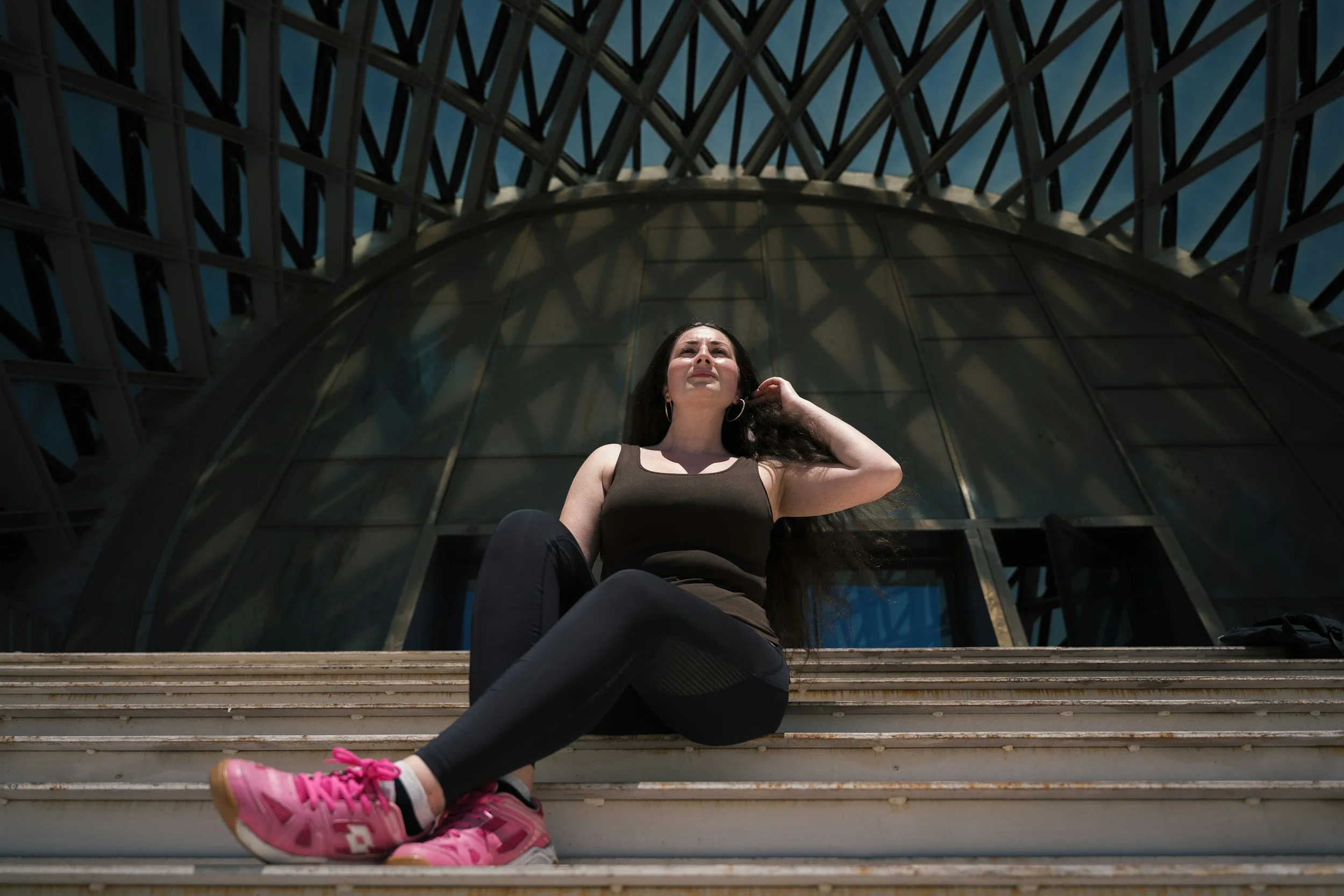 Young woman with long dark hair sitting on steps outside, looking up with a thoughtful expression, against the background of a modern glass and steel architectural structure.