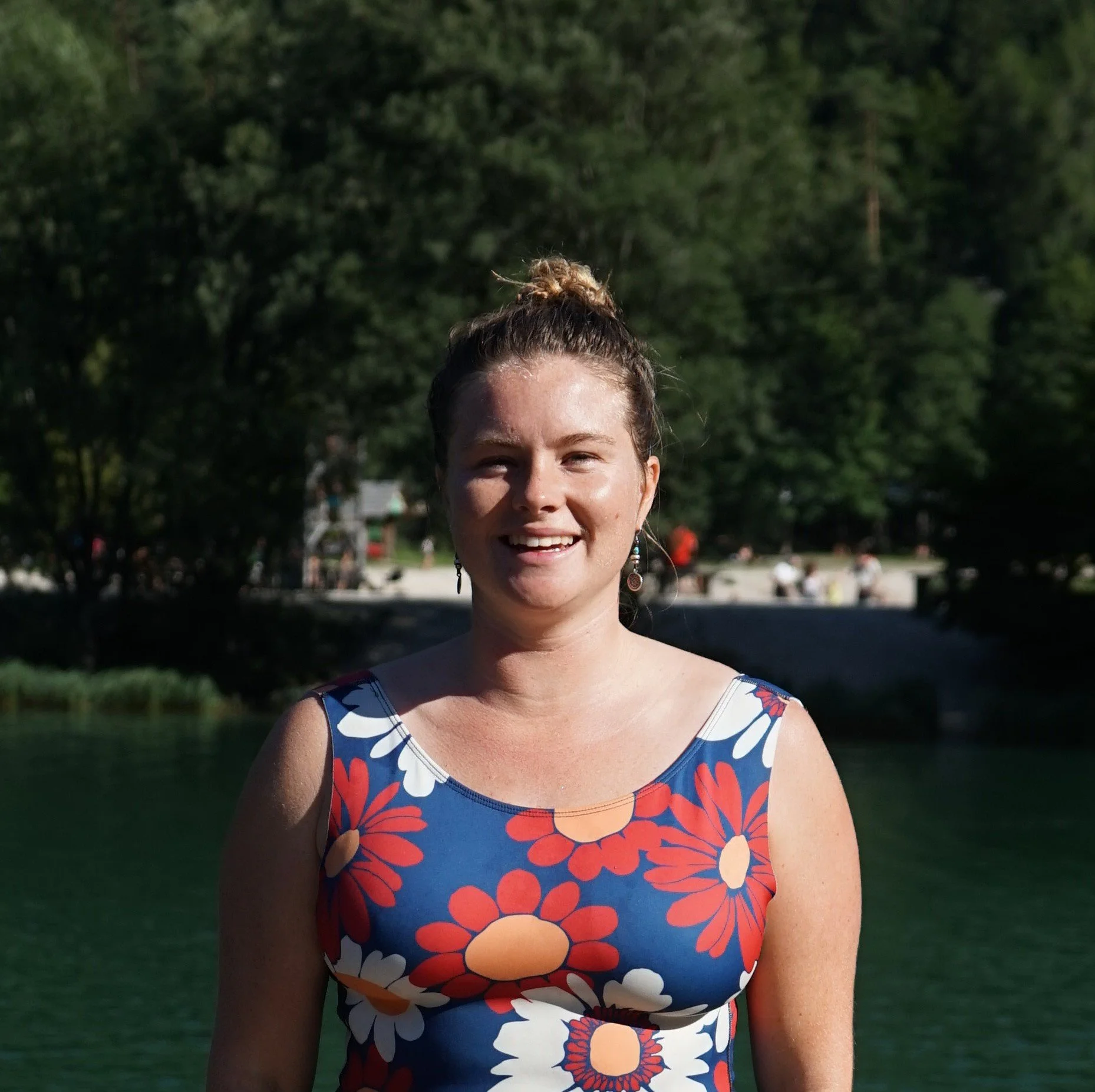 A woman smiling outdoors near a body of water, wearing a blue dress with a floral pattern in red, white, and orange, with green trees in the background.