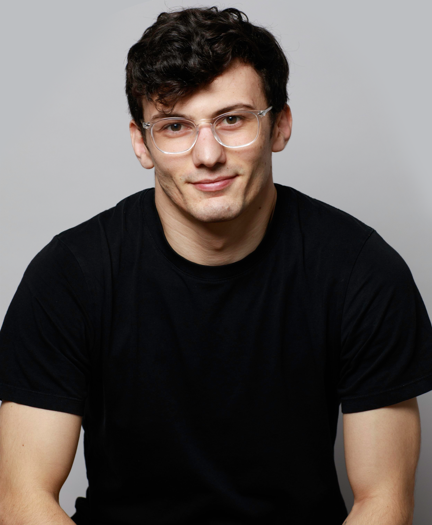 A young man with dark curly hair wearing clear glasses and a black t-shirt, sitting against a plain gray background.