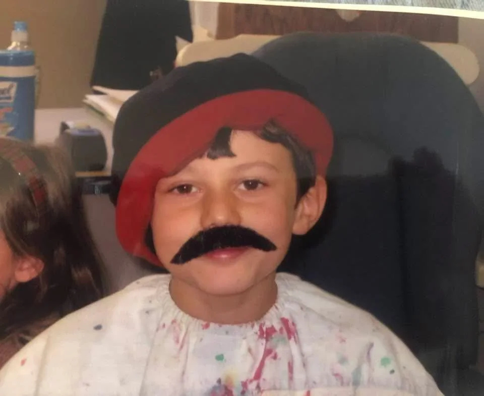 Child with a paper fake mustache wearing a red beret, sitting in a chair.