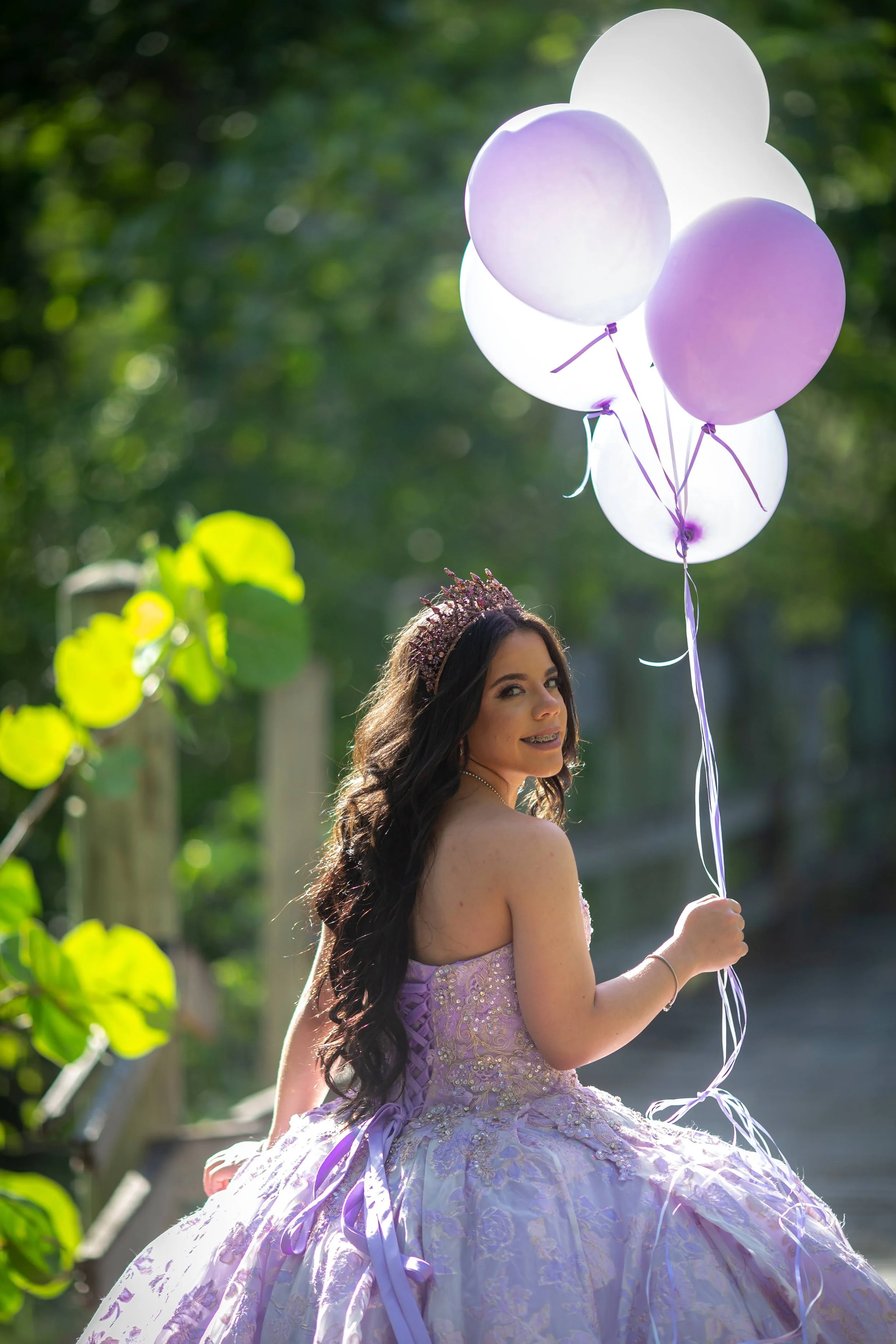 Young woman in a lavender ball gown and tiara holding purple and white balloons outdoors on a sunny day.