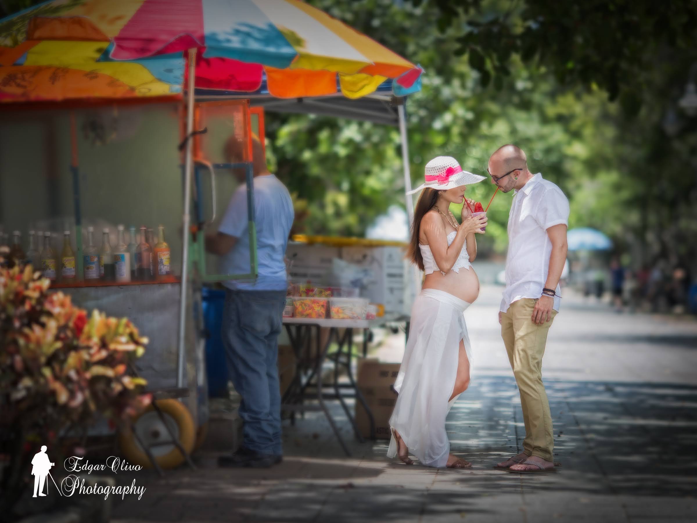 A man and a pregnant woman are standing close together, sharing a drink with red straws during daytime outdoors near a water body. The woman is wearing a wide-brimmed sun hat with a pink ribbon and a white dress, and the man is wearing glasses, a whi