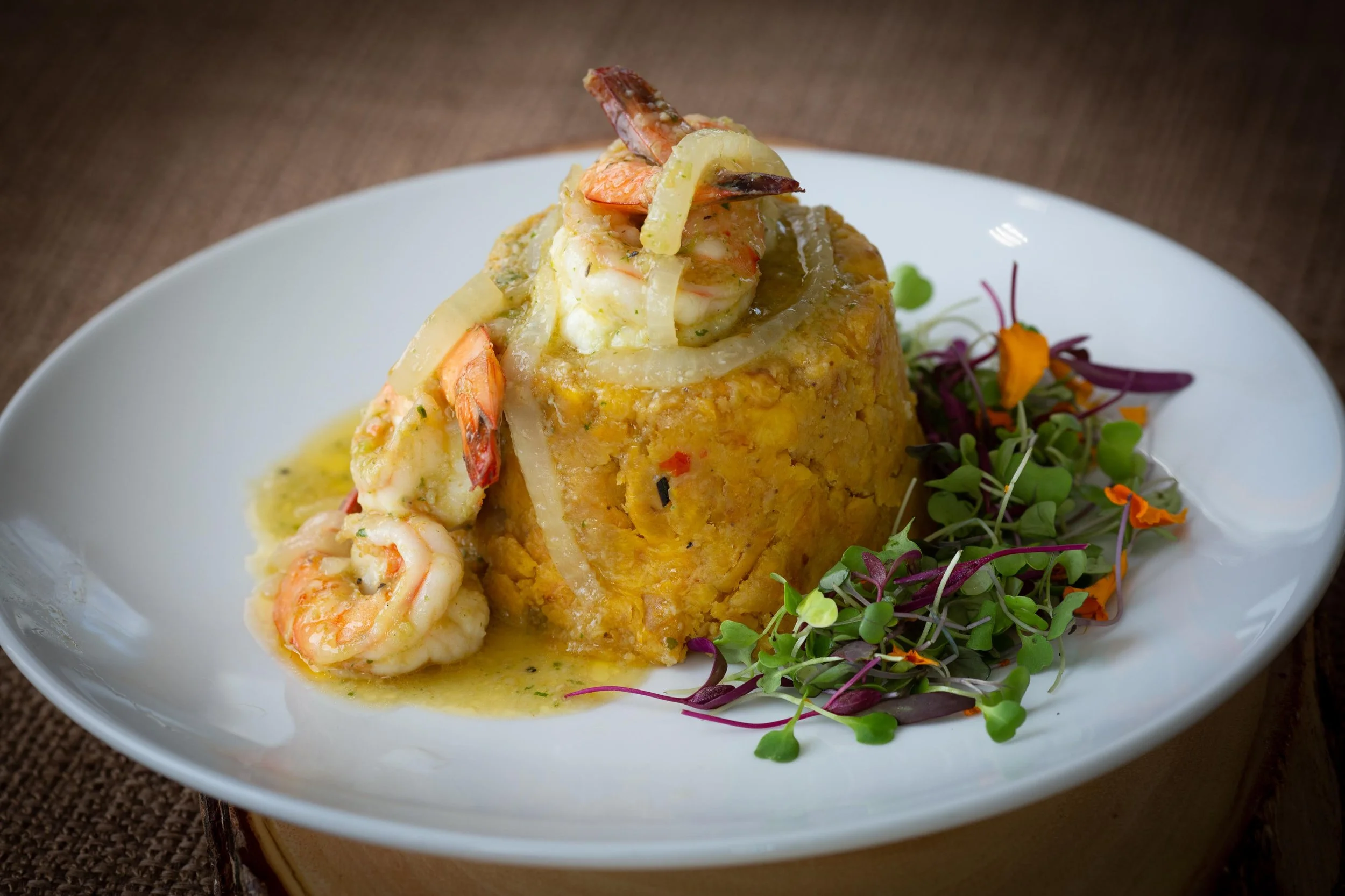 A plated dish with a large portion of cornbread topped with shrimp and sauce, with a side of microgreens and colorful edible flowers.