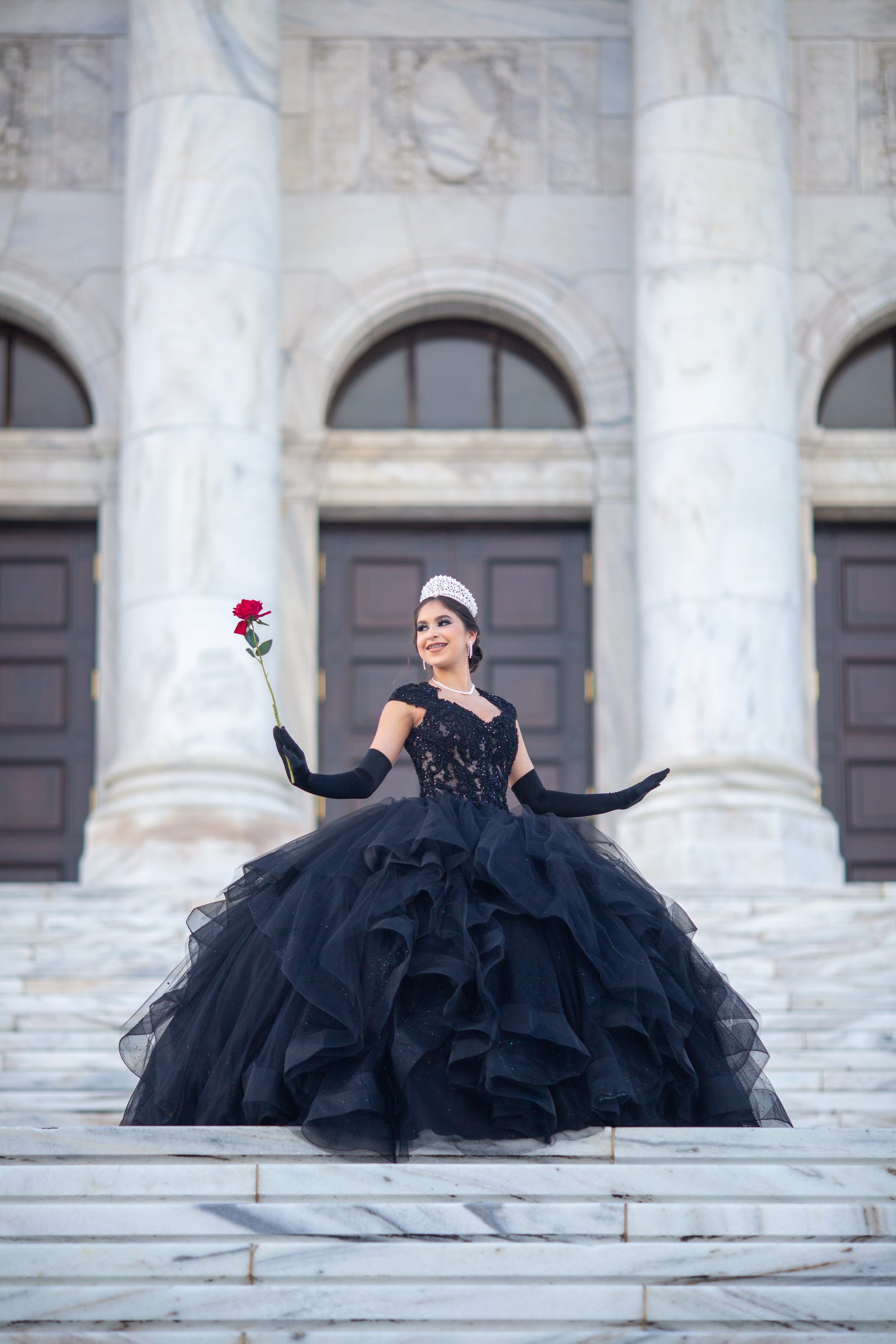 A woman in an elegant black gown and tiara, holding a red rose, stands on marble stairs in front of a building with large columns and arched windows.