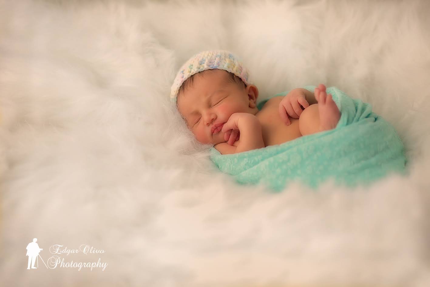 A peacefully sleeping newborn baby wrapped in a light blue blanket, lying on a soft white fur surface, wearing a pastel-colored knit hat.