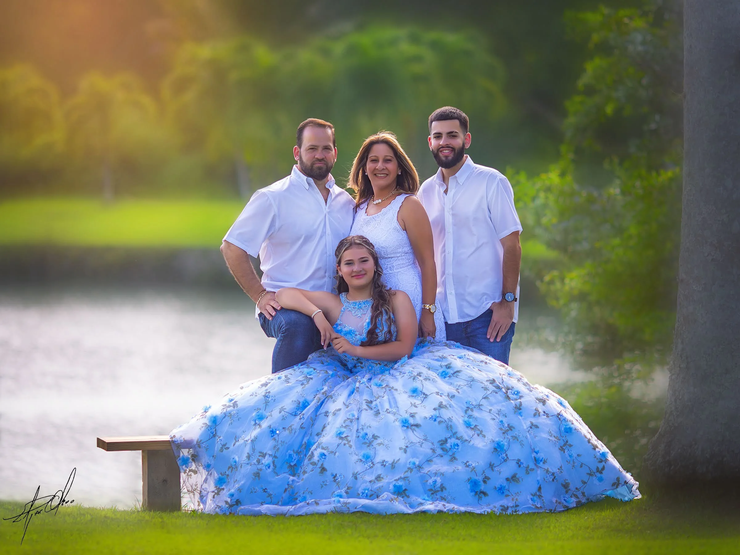 A family of five stands outdoors near a body of water, with trees in the background. The woman is wearing a white dress, and the three men are dressed in white shirts and blue jeans. The young girl is dressed in a blue floral dress, sitting on a wood