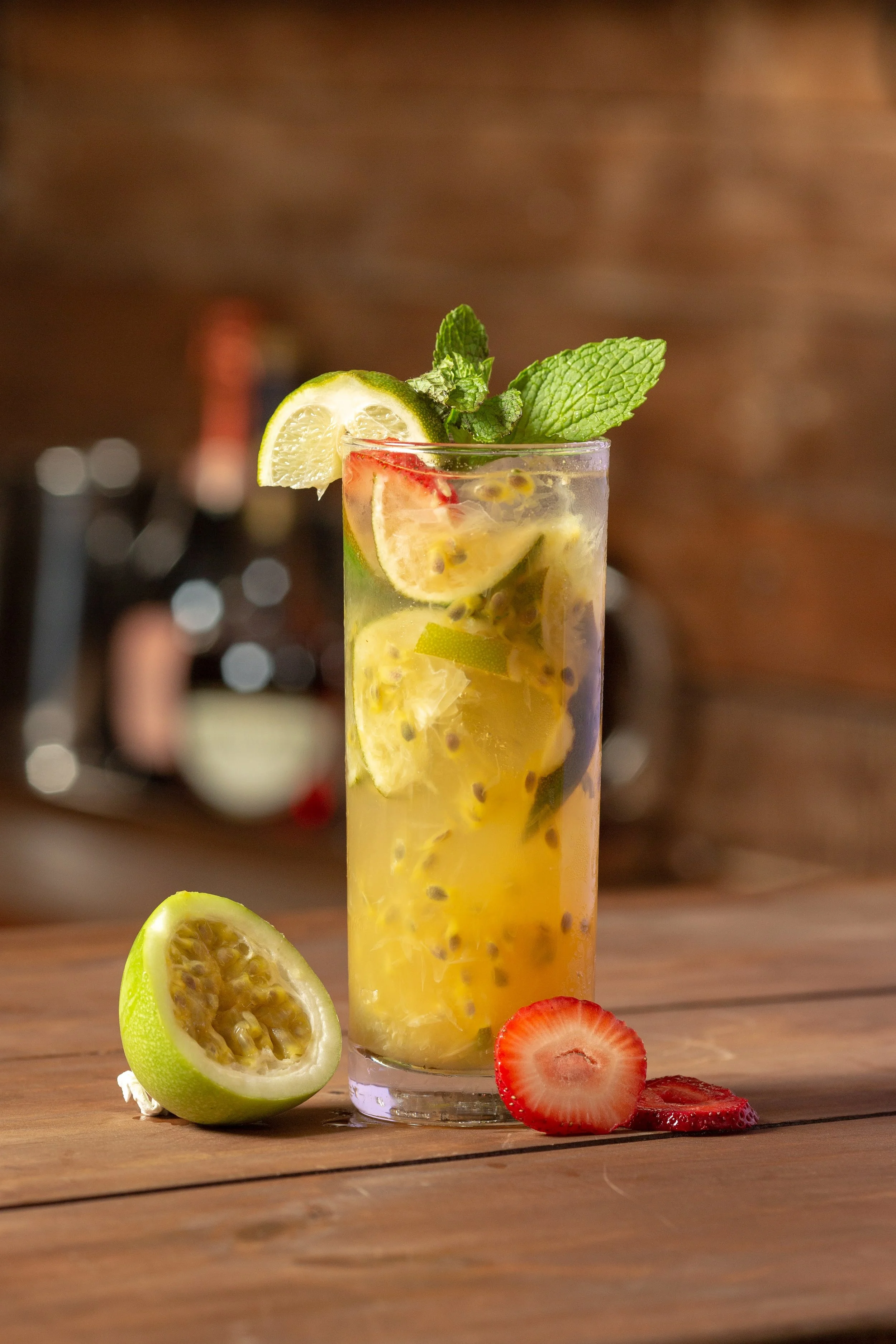 A tall glass of passion fruit mojito garnished with a lemon wedge and mint leaves, with passion fruit seeds floating inside. Fresh strawberries and a halved passion fruit are placed on the wooden table beside the glass, set against a blurred backgrou