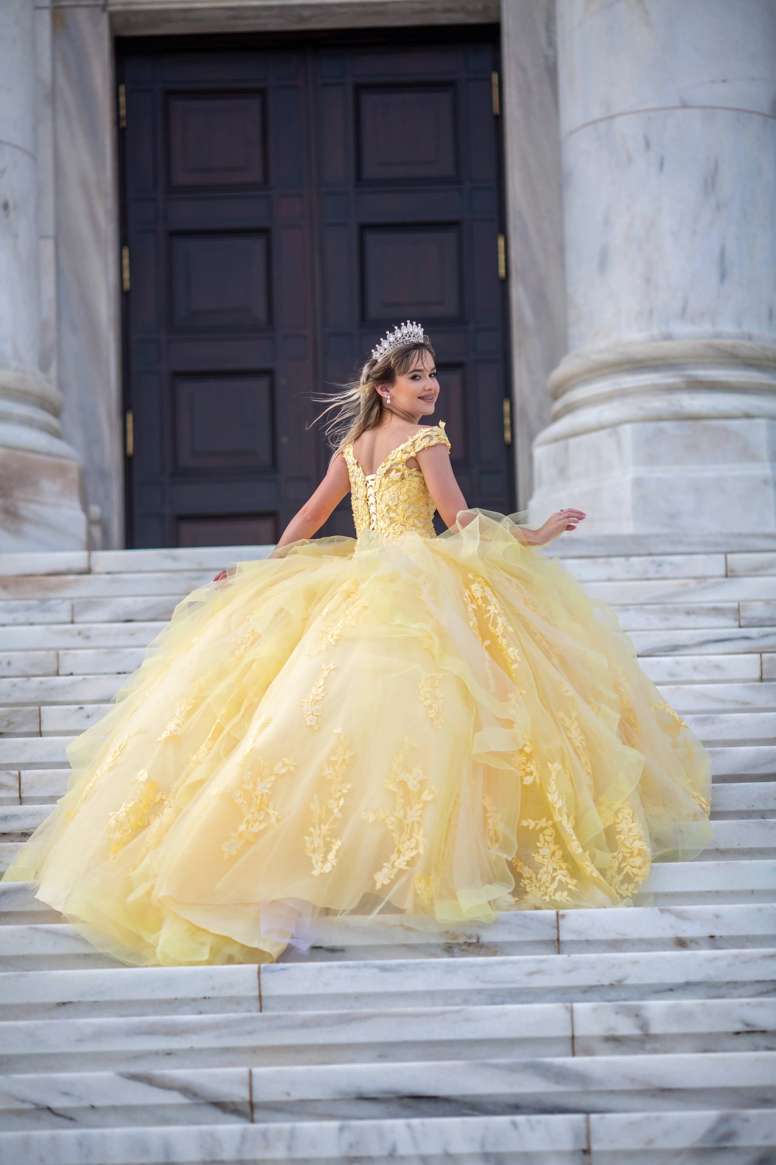A young woman in a yellow ball gown and tiara sitting on marble stairs in front of large dark wooden doors.
