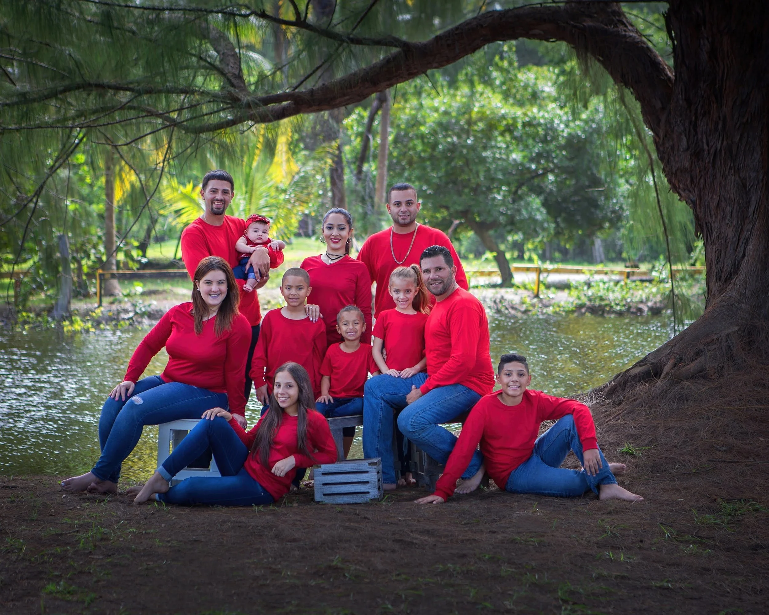 A large group of people, including children and adults, standing and sitting outdoors on a dock by a creek in a wooded area, all wearing red shirts.