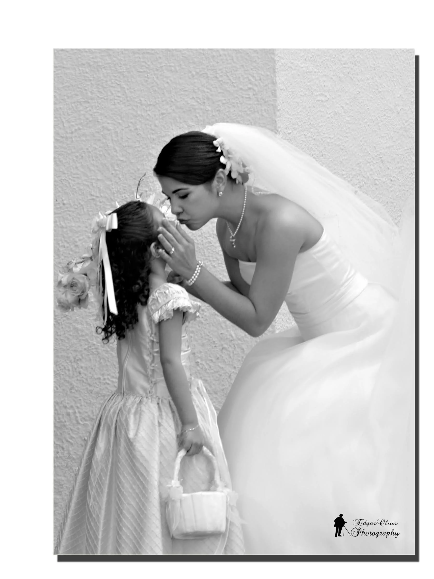 Black and white photo of a bride and a young girl, likely a flower girl, sharing a tender moment on her wedding day. The bride is leaning down and gently kissing the girl. The bride is wearing a wedding dress with a veil decorated with flowers, and t