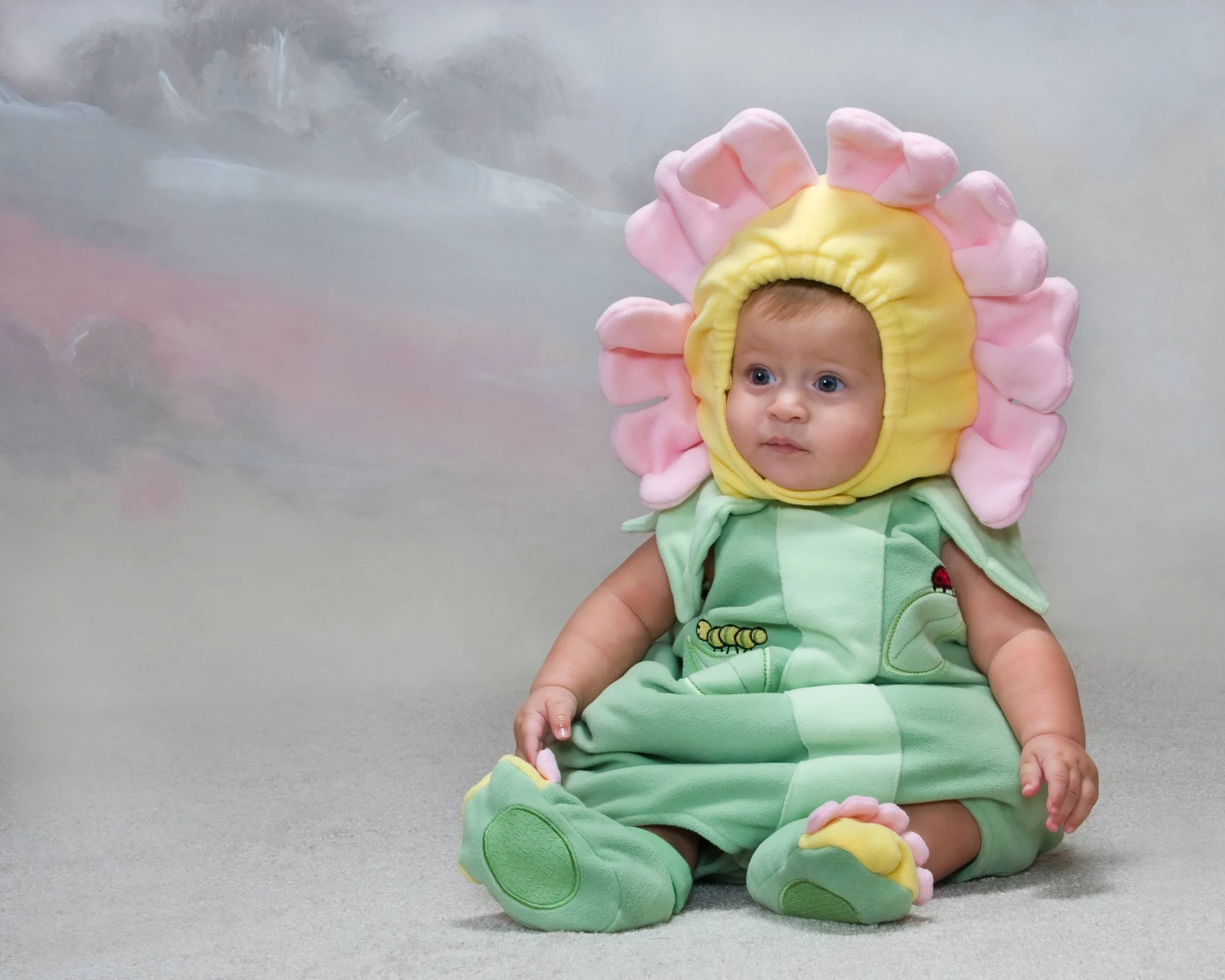 A baby dressed in a lion costume with pink and yellow mane, green body, and lion paw shoes, sitting on a gray surface.