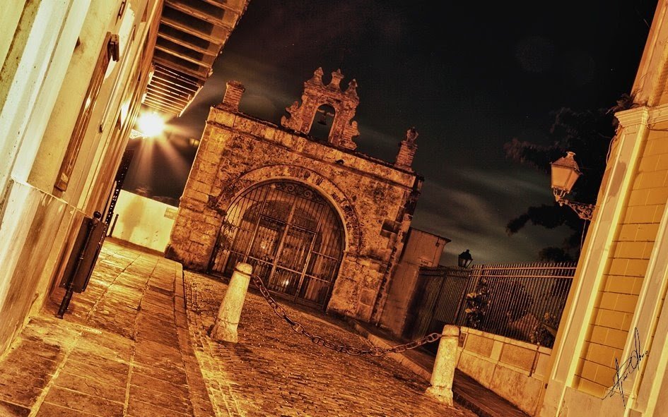 Nighttime view of an old stone gate with metal bars, surrounded by historic stone and brick buildings, illuminated by streetlights.