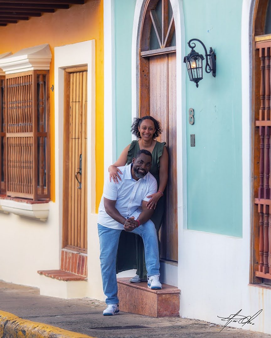 A smiling woman and man pose in front of a colorful house entrance, with the woman standing behind the man who is leaning on the doorstep.