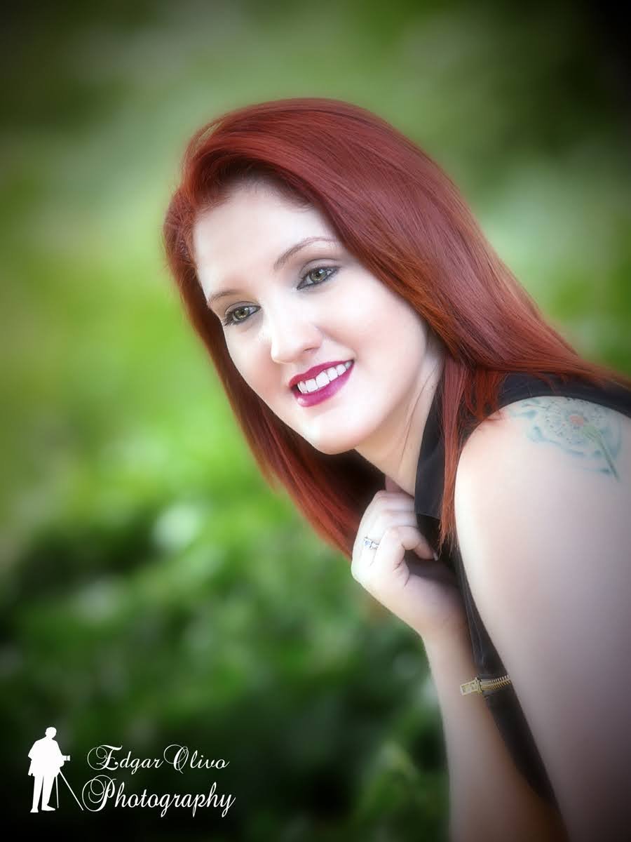 A young woman with red hair and makeup, smiling outdoors with a green blurred background.