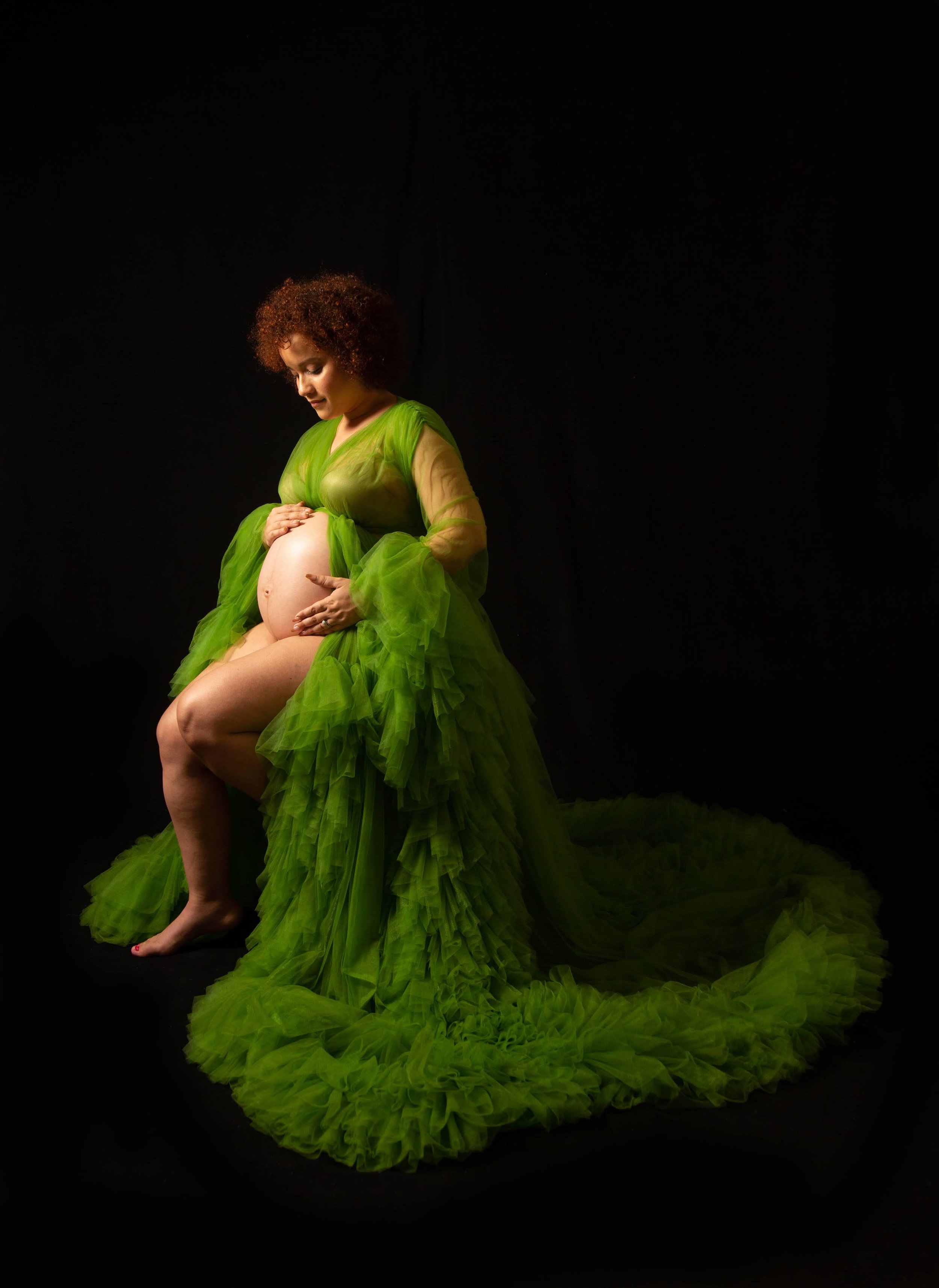 A pregnant woman with red curly hair, wearing a flowing bright green dress, sitting with her legs crossed and gently touching her belly, against a dark background.