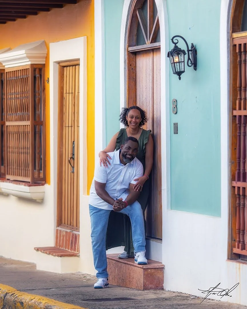 Smiling woman and man in white shirt and jeans posing in front of colorful houses with wooden doors and windows, and a black lantern outside.