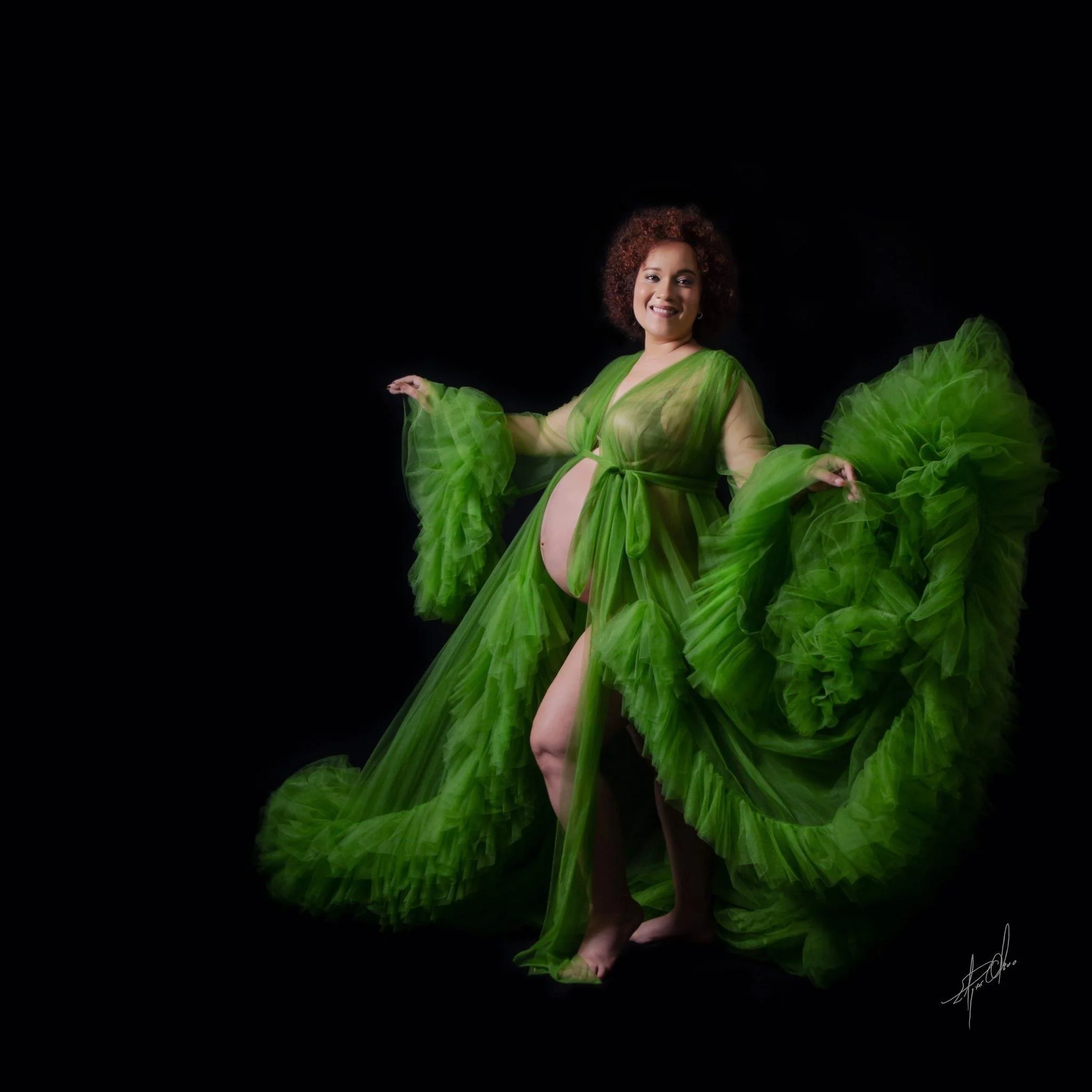 A woman with curly hair wearing a flowing green dress, standing against a black background, smiling with her arms extended, surrounded by flowing green fabric.