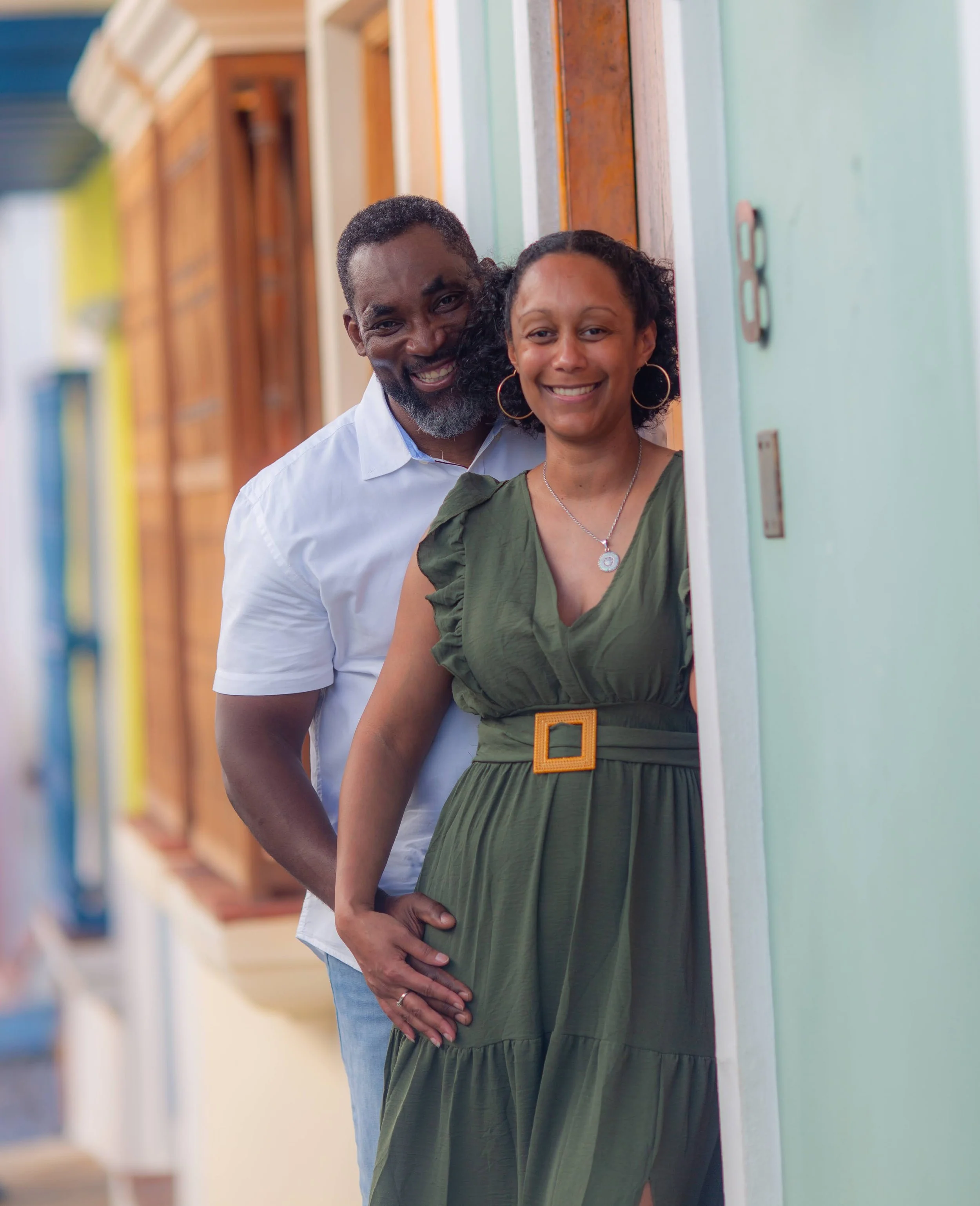 A couple is standing in a hallway, with the man touching the pregnant woman’s belly. Both are smiling and looking at the camera.