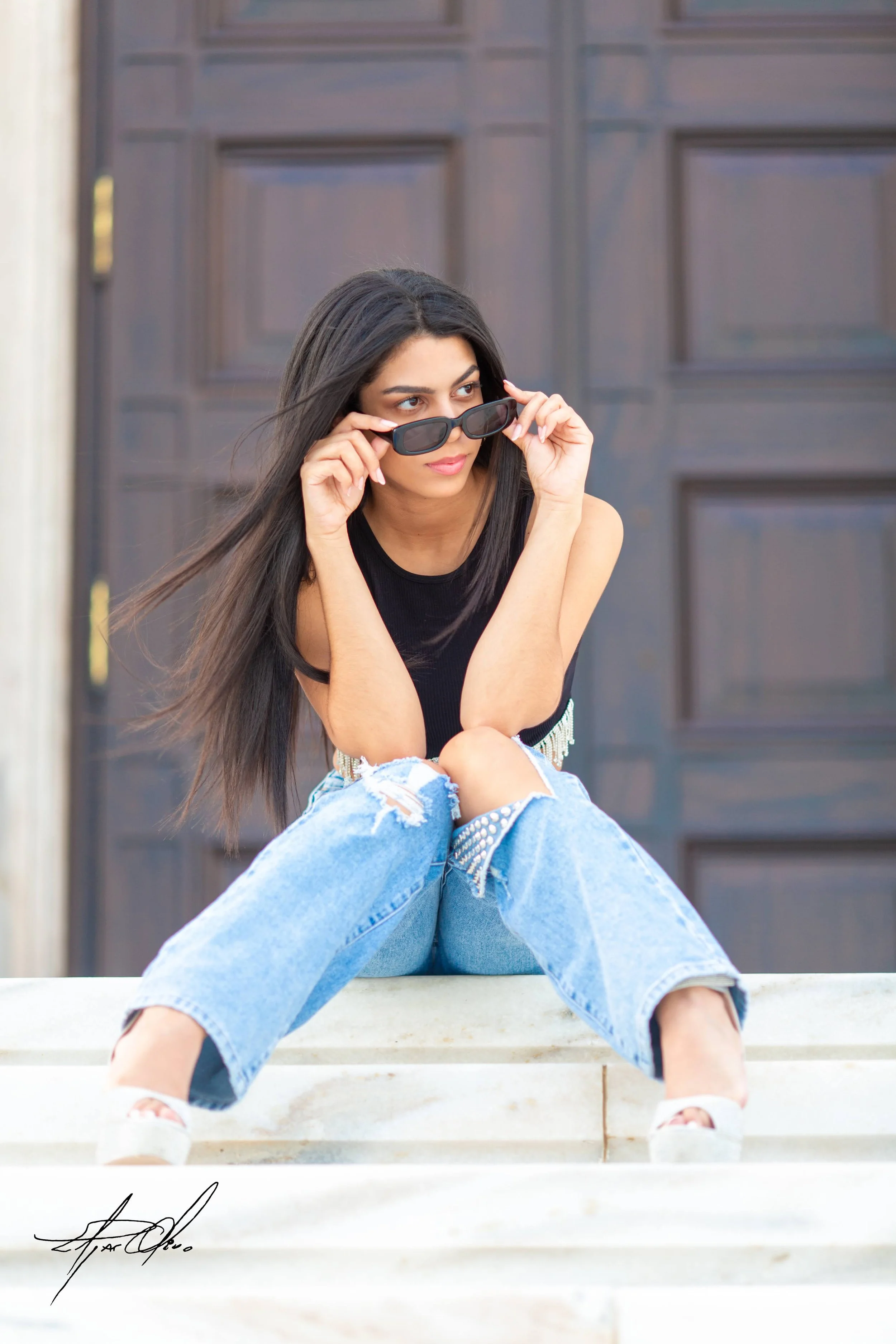 A young woman with long dark hair, wearing a black sleeveless top and ripped blue jeans, sitting on concrete steps in front of a dark brown wooden door, holding sunglasses and looking to the side.