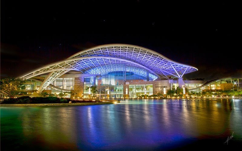 Night view of a modern building with a curved, illuminated roof reflecting on a body of water.