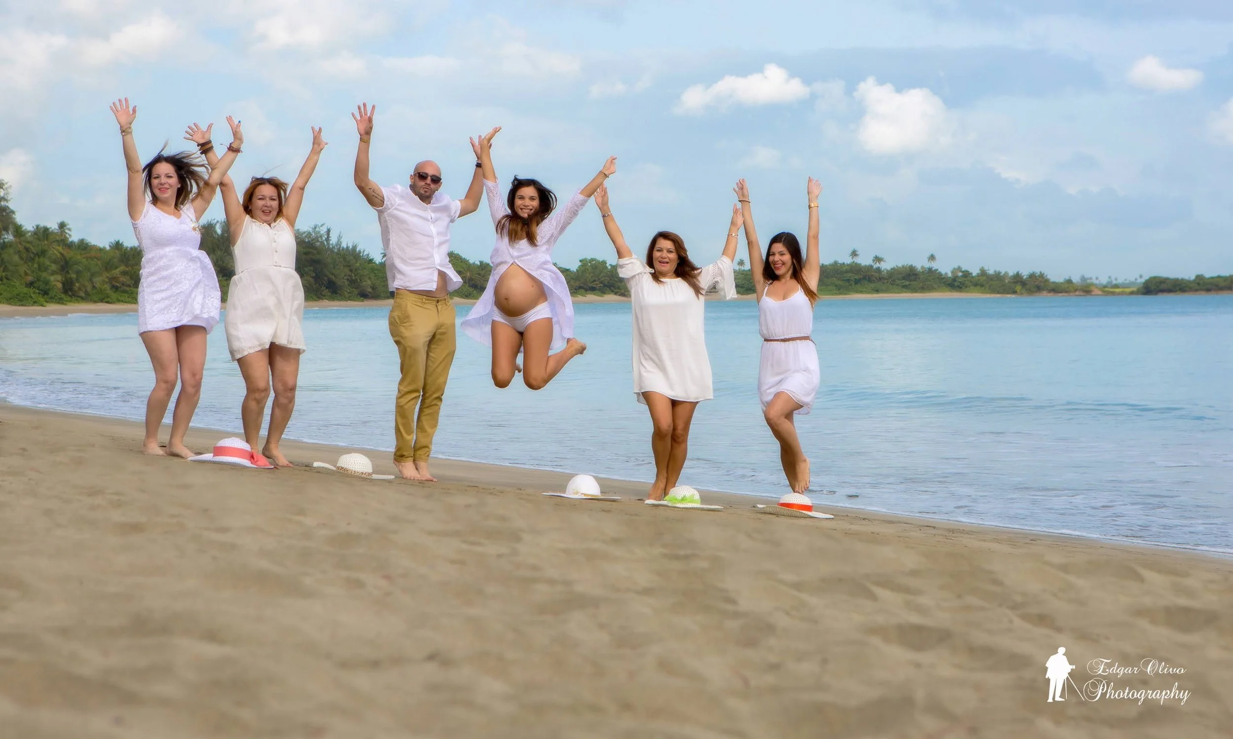 A group of six people, including pregnant women and men, joyfully jumping on a sandy beach near the water, wearing casual white outfits.