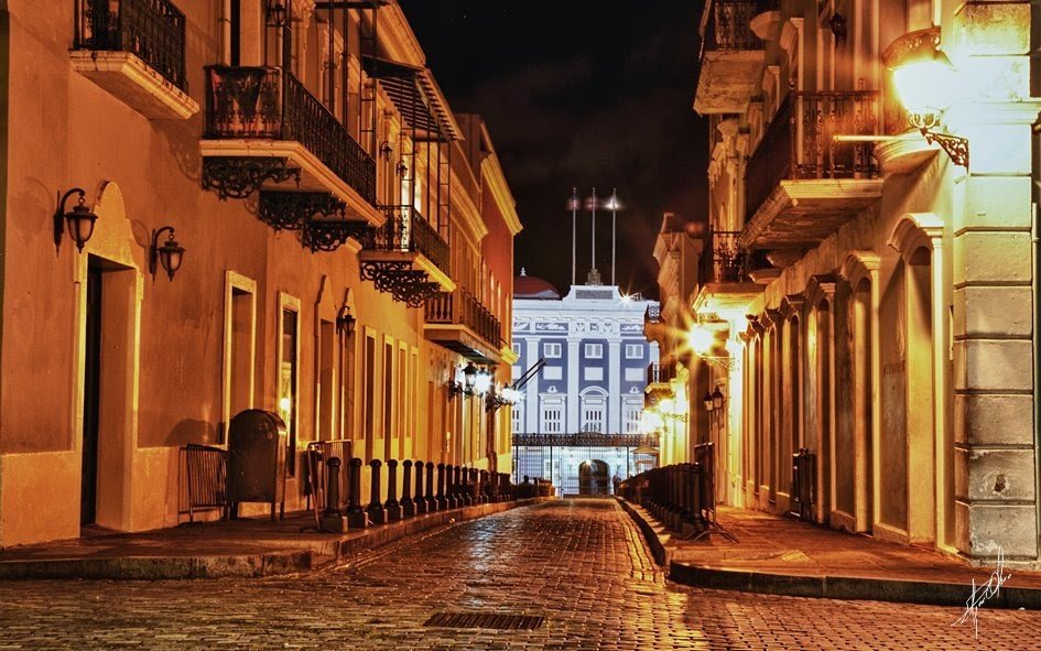 Nighttime view of a quiet, cobblestone street lined with historic buildings, illuminated by streetlights, with an ornate building in the background.