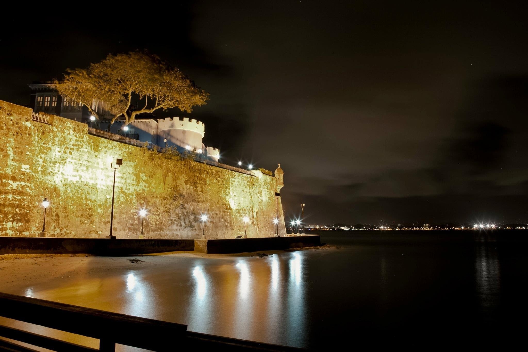 Nighttime view of a historic stone fortress or city wall with bright lighting, a large tree, and a calm body of water reflecting the lights, with a dark cloudy sky and city lights in the distance.