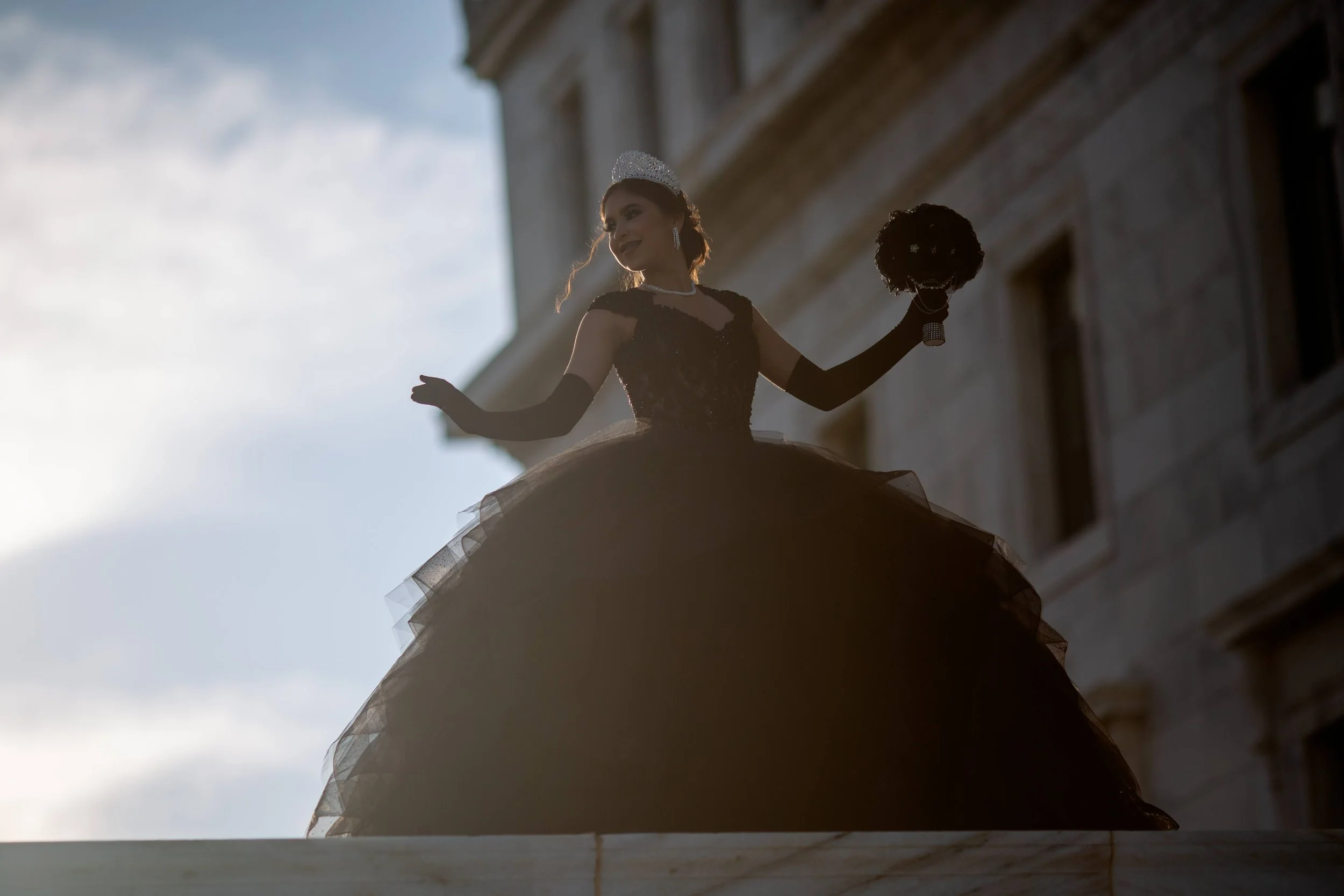 A woman dressed in a formal black gown with a tiara, holding a bouquet, standing on a balcony or ledge outside a building, with the sky and part of the building visible behind her.