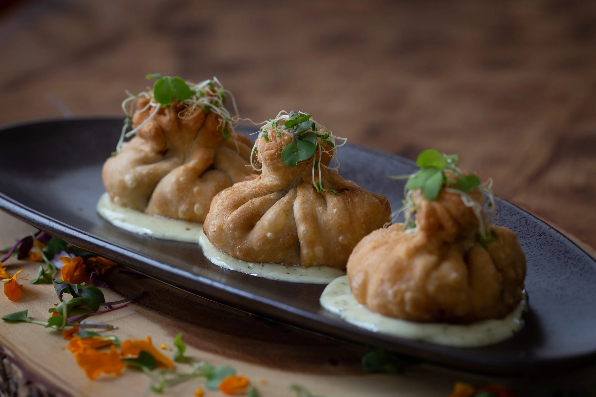Close-up of Indian vegetable dumplings called momos on a black plate, garnished with microgreens, served with green chutney, and decorated with orange flower petals.