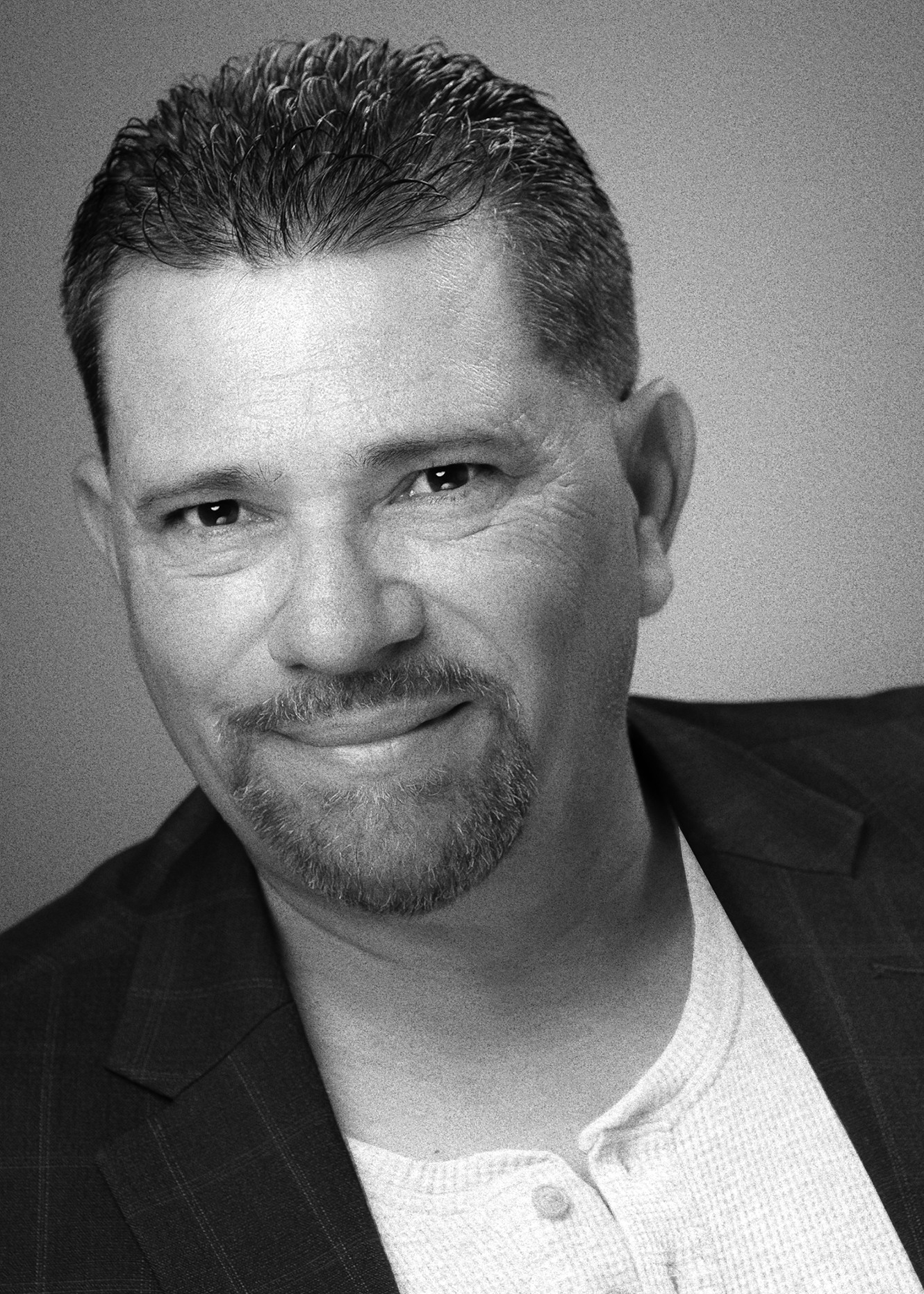 Black and white portrait of a smiling man with short, combed-back hair, a trimmed beard, and wearing a plaid blazer over a light-colored sweater and shirt.