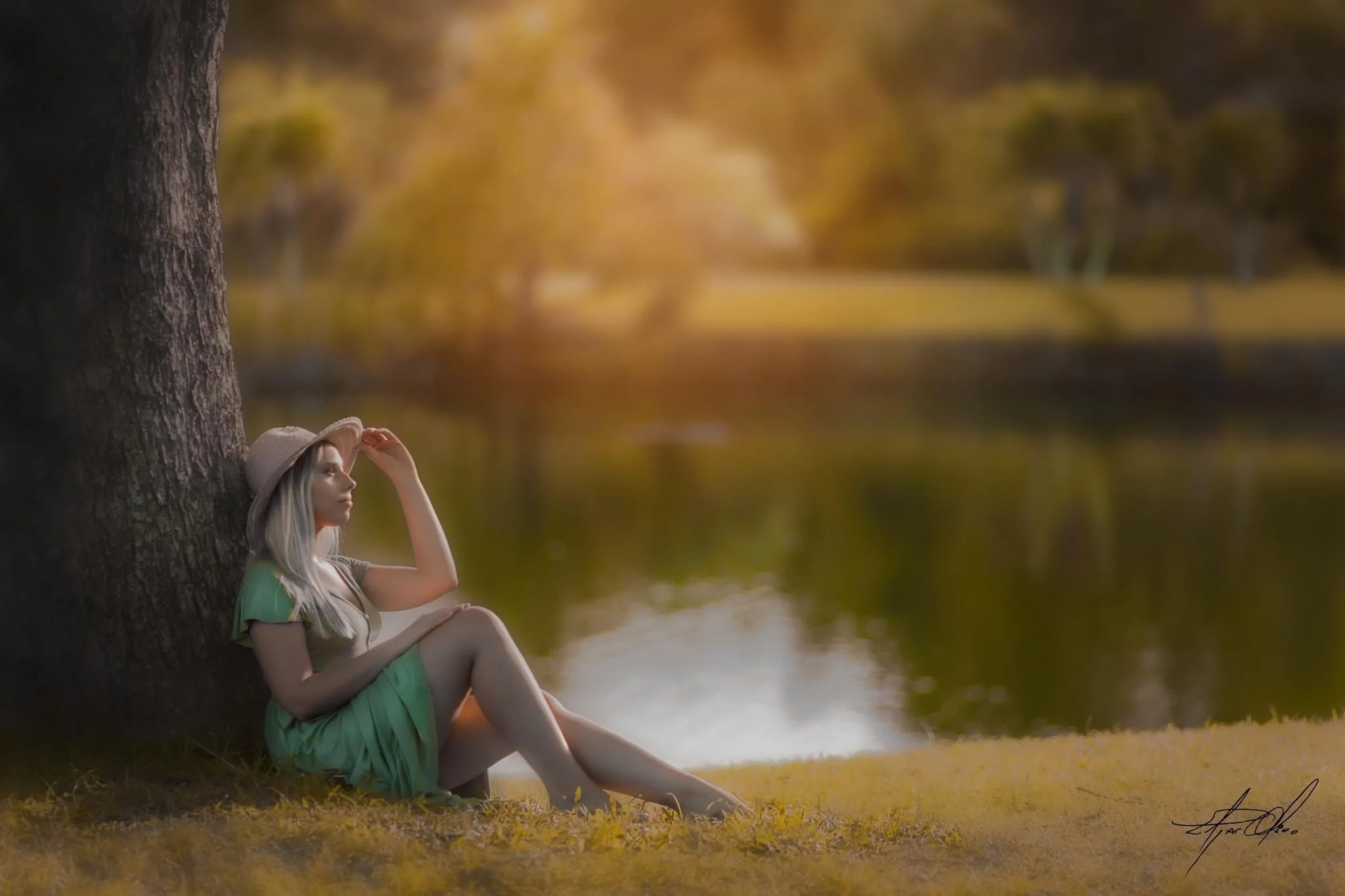 A woman with long blonde hair, wearing a beige hat, a green skirt, and a beige top, is sitting and leaning against a large tree near a calm lake during autumn. She is holding her hat with her right hand and gazing thoughtfully into the distance.