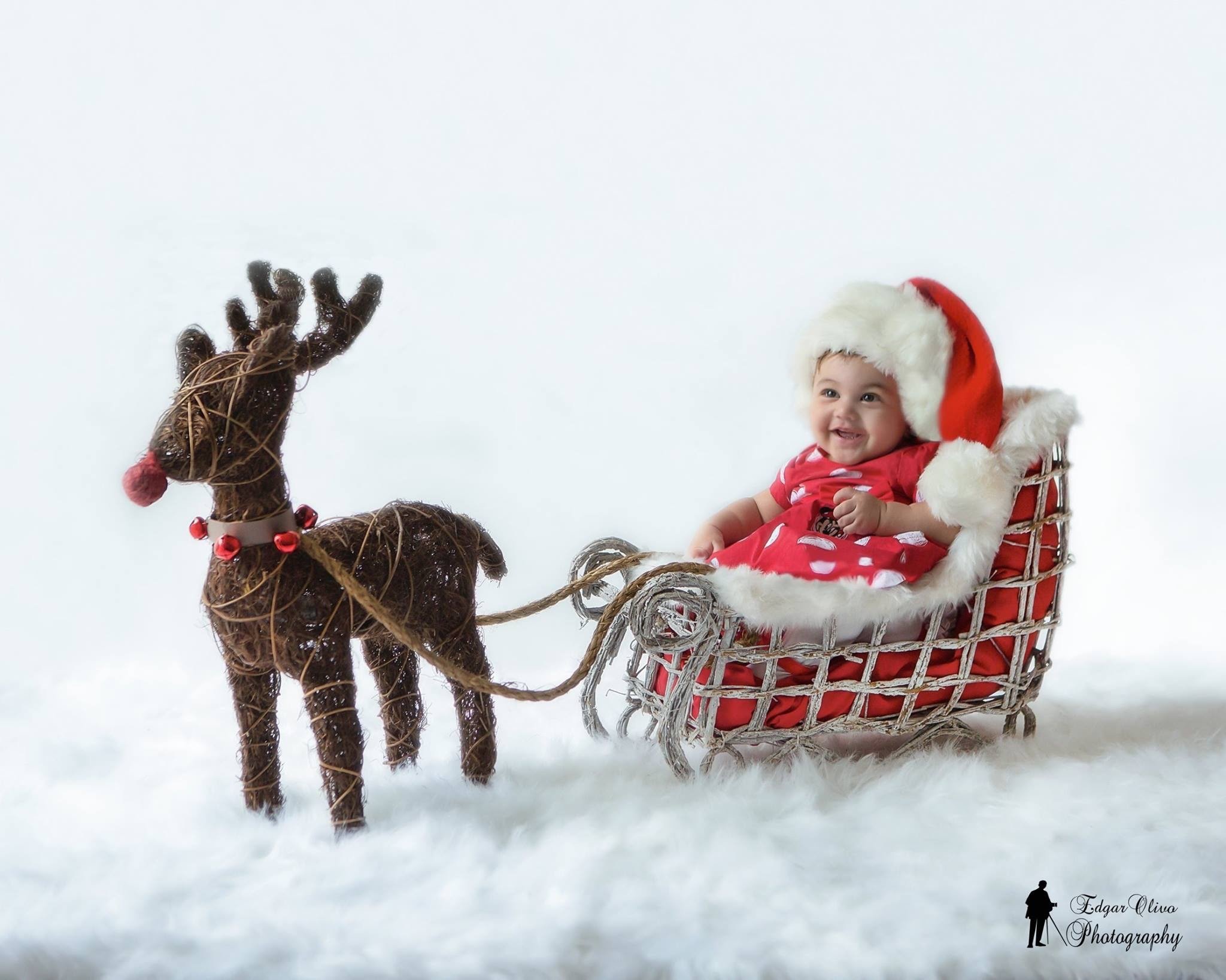 A baby with a Santa hat smiling while seated in a red sled being pulled by a reindeer figurine on snowy ground.