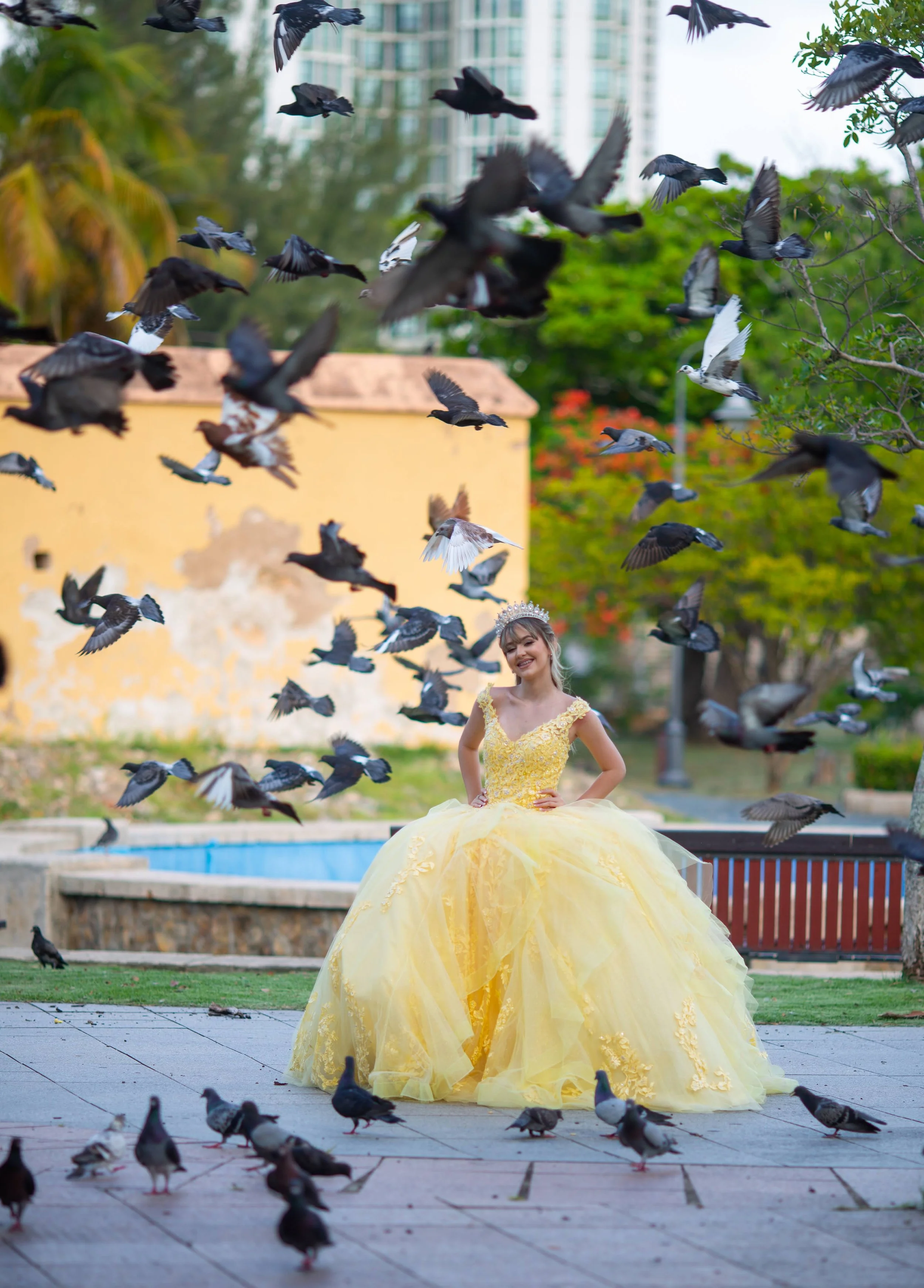 A young woman in a yellow ball gown and tiara standing smiling among a flock of pigeons in an outdoor park with trees, buildings, and a fountain in the background.