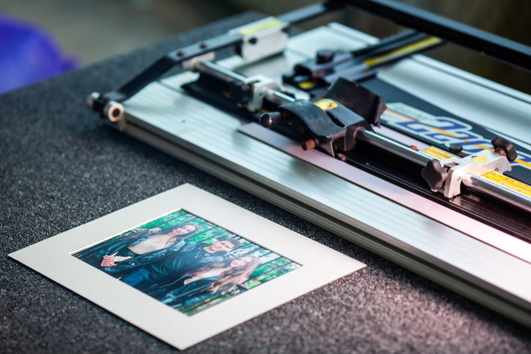 Photograph of a photo printing machine with photographs being produced on photo paper, featuring a woman in black clothing making a peace sign with her hand.