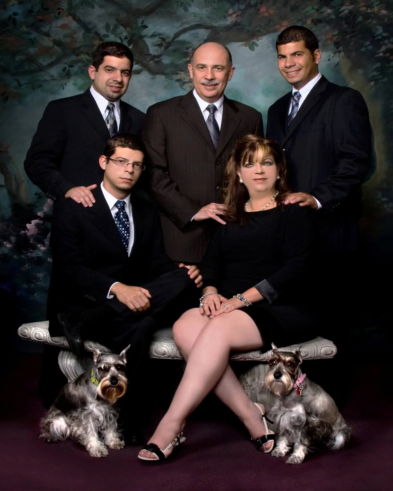 Group portrait of a man with four younger adults, two men and one woman, and two dogs, on a decorative bench in front of a painted backdrop.