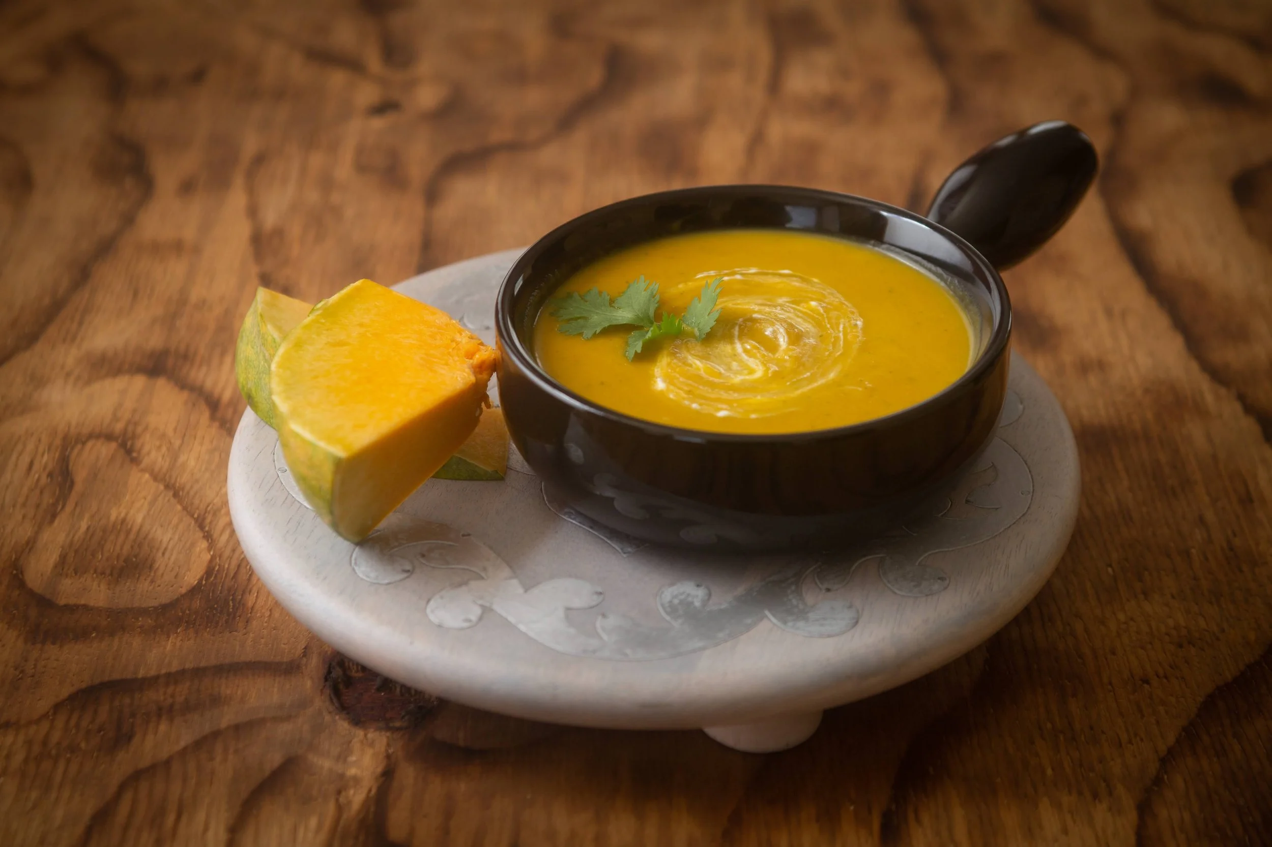 A bowl of bright orange pumpkin soup garnished with a cilantro leaf, served with a wedge of pumpkin on a white plate, placed on a rustic wooden table.