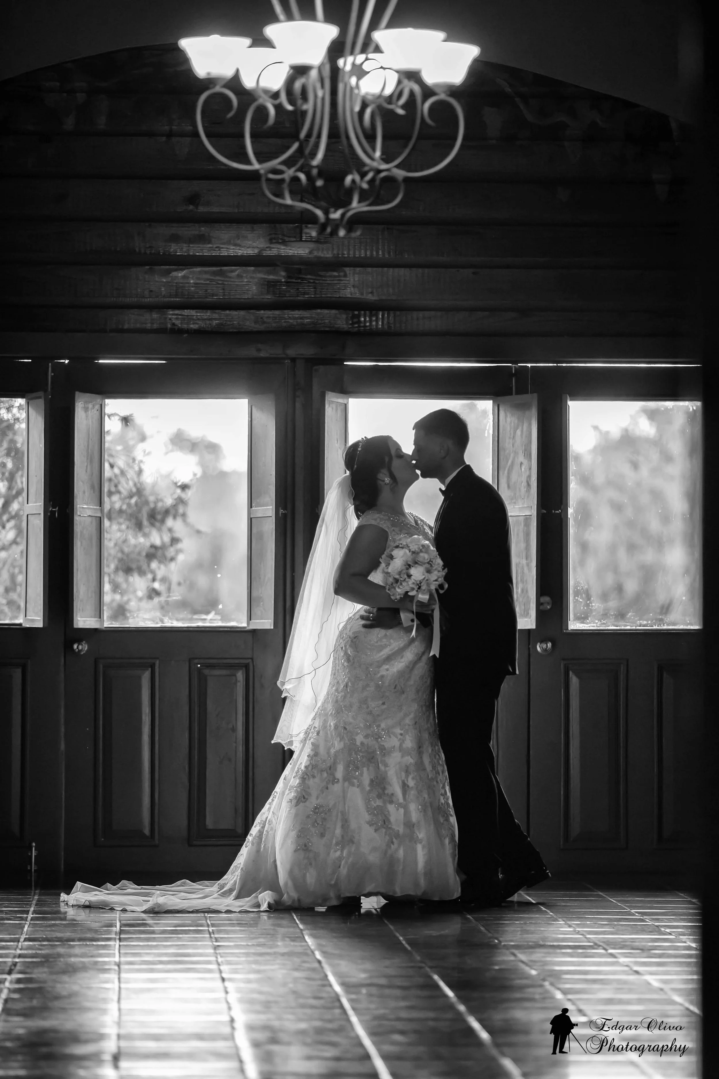 A bride and groom sharing a kiss in front of open windows with light streaming in. The bride is wearing a lace wedding dress and holding a bouquet, and the groom is in a suit. There is a chandelier hanging above them.