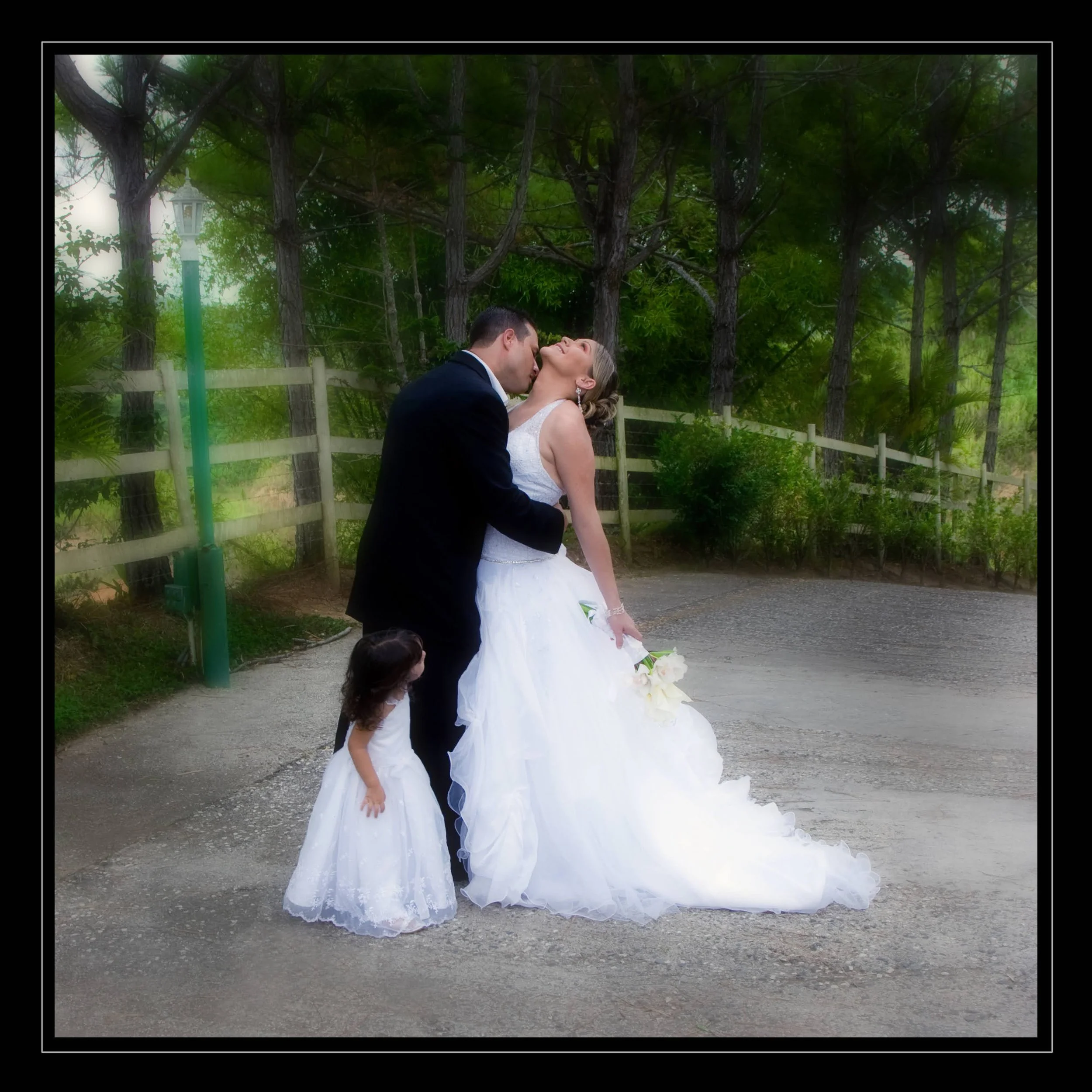 A bride and groom sharing a kiss outdoors amid trees, with two young girls in white dresses nearby.