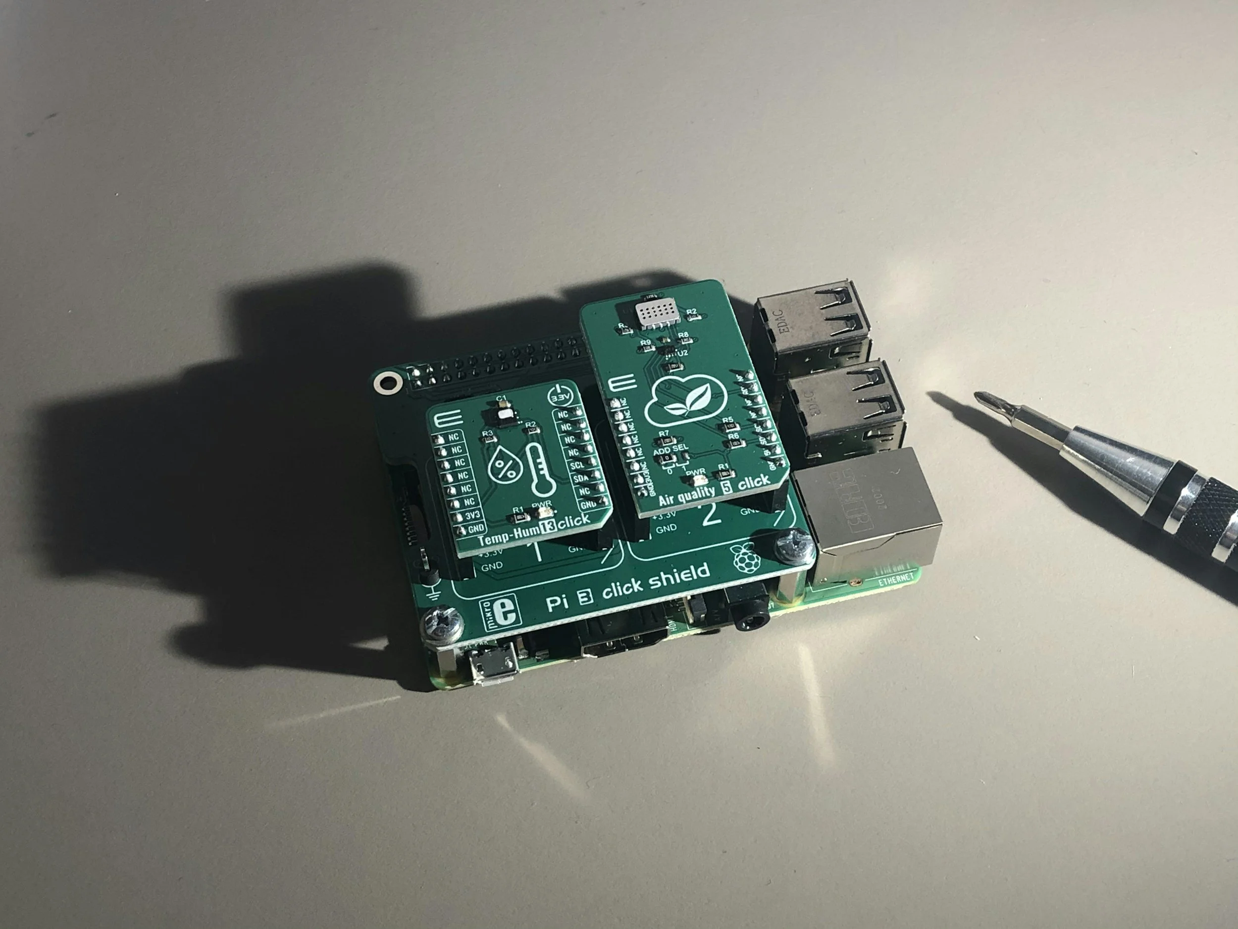 A close-up of a green Raspberry Pi 3 Click Shield with various electronic components and ports on a gray surface, with a screwdriver nearby.