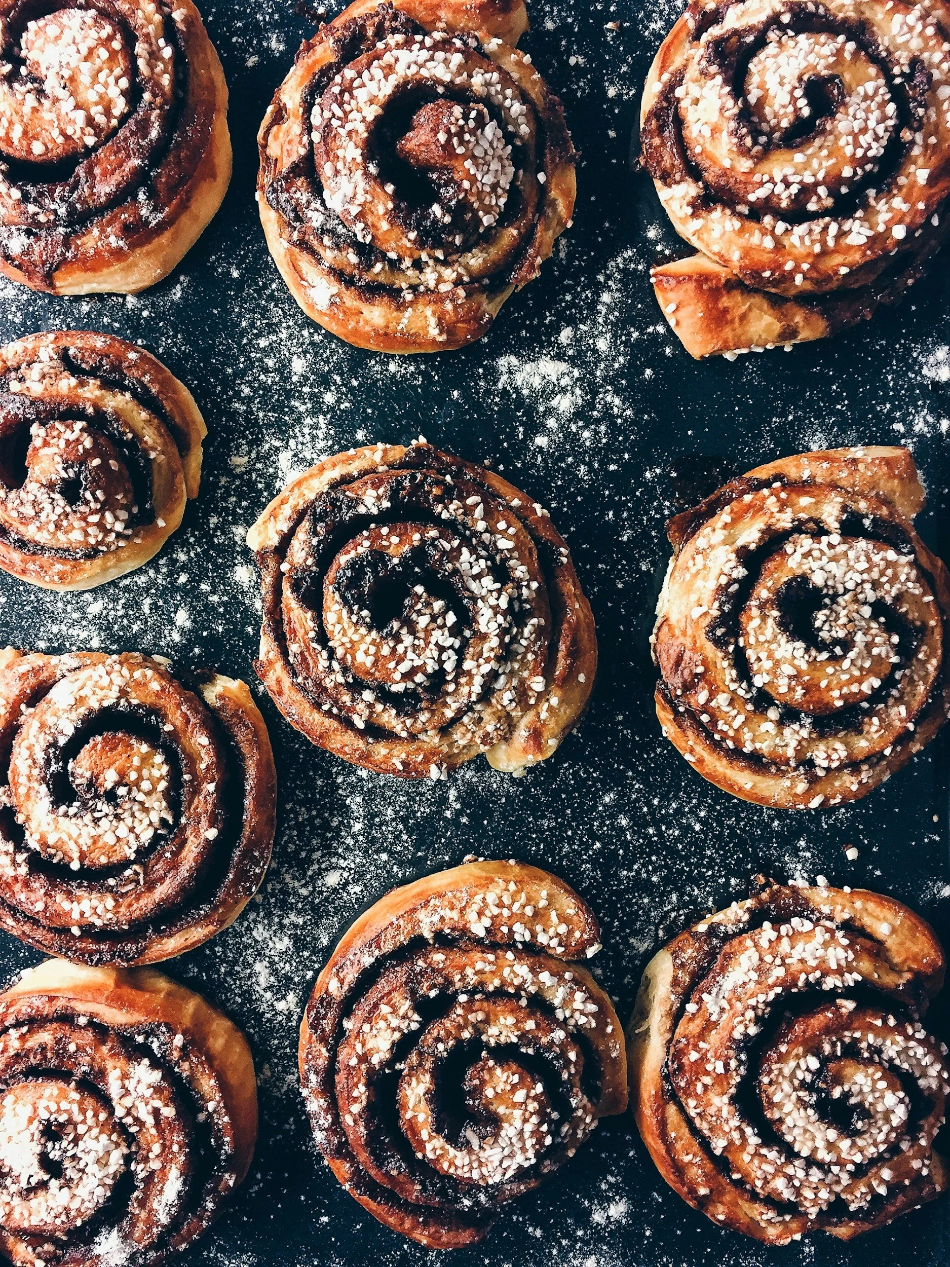 Several cinnamon rolls topped with icing sugar and chocolate drizzle on a black surface.