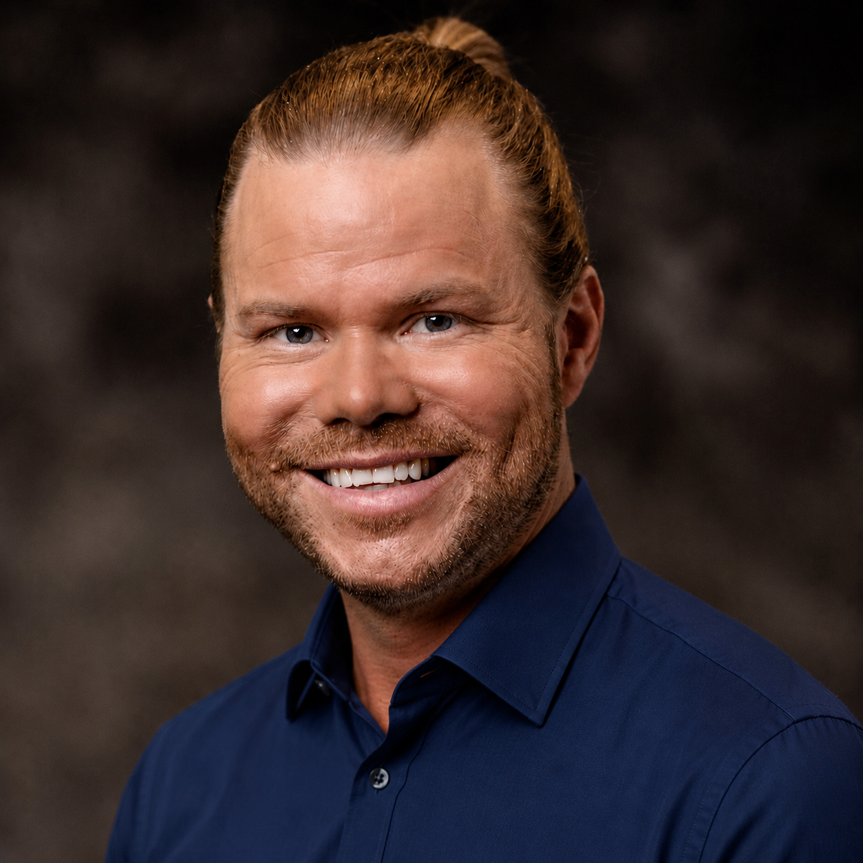 Headshot of a smiling man with red hair, a beard, and blue eyes, wearing a navy blue shirt, against a dark background.