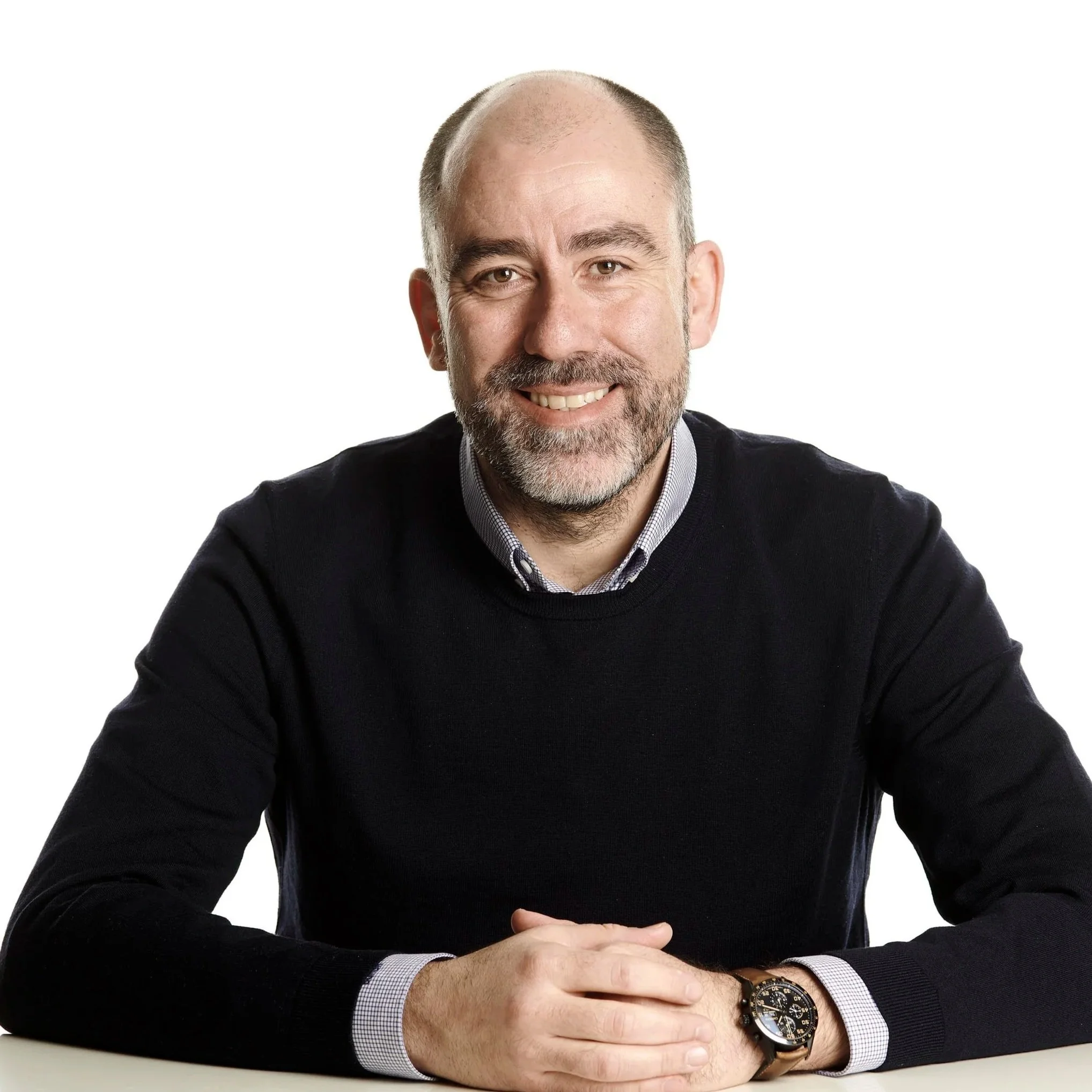 Retrato de un hombre con barba, sonriendo, vestimenta formal con camisa de cuadros y suéter negro, usando reloj de pulsera, frente a fondo blanco.